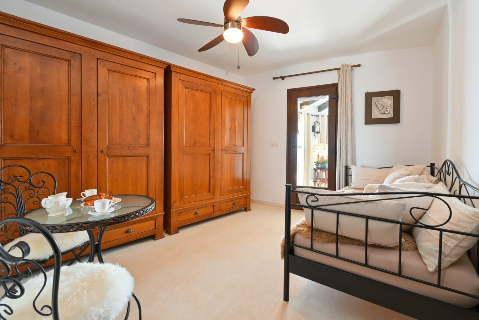 Bedroom featuring two large rustic wooden wardrobes, a black wrought iron daybed, and a bistro table set. Includes a wooden ceiling fan and glass door access.