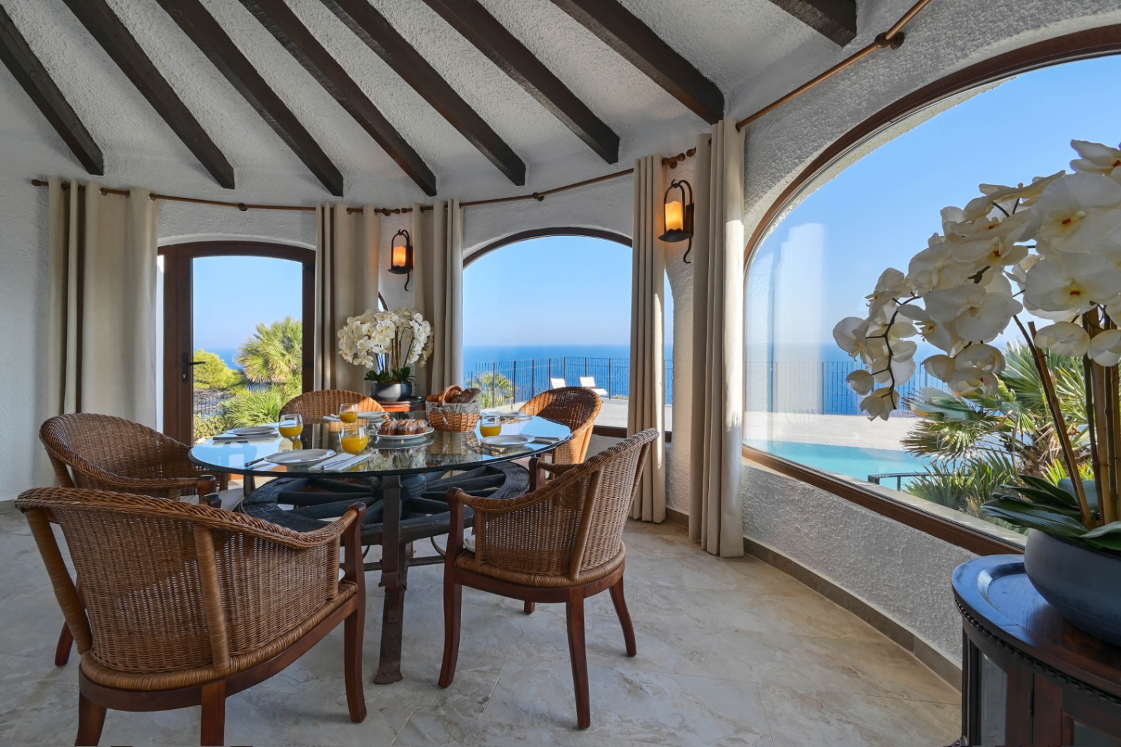 Circular dining area featuring a wagon-wheel glass table and wicker chairs, set under an exposed beam ceiling with panoramic sea and pool views through arched windows.