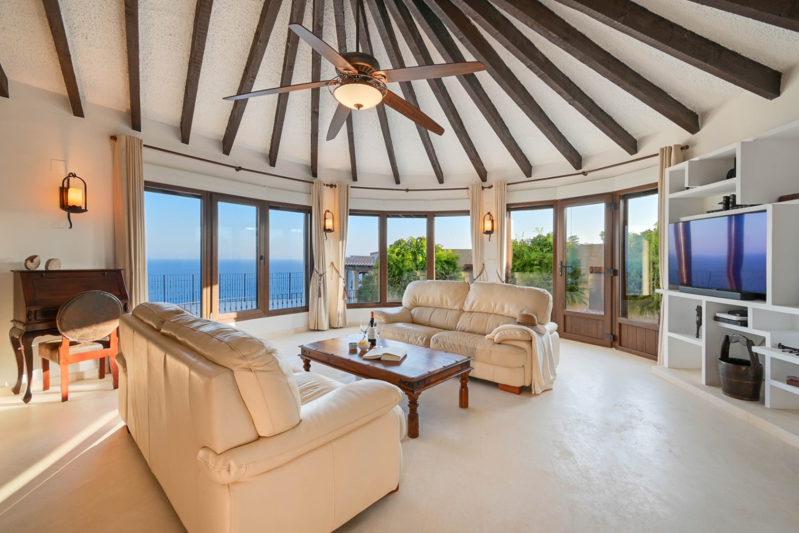 Circular living room featuring a vaulted ceiling with dark wooden beams, leather sofas, and panoramic sea views through large windows.