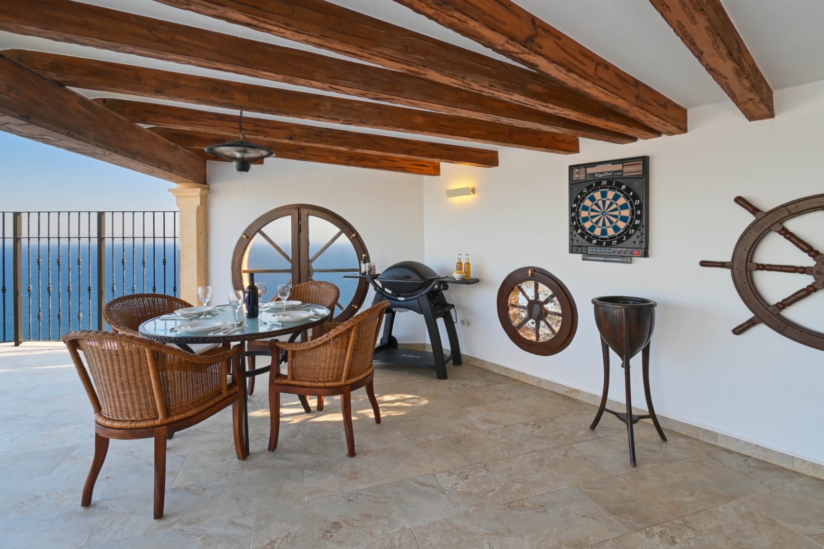 Covered terrace featuring exposed wooden beams, circular porthole windows, and sea views. Includes a wicker dining set, gas grill, and nautical wall decor.