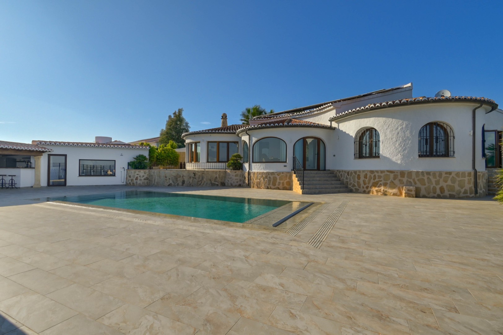 Exterior view of a Mediterranean villa featuring a rectangular swimming pool, stone-clad walls, and a spacious tiled terrace under a clear sky.