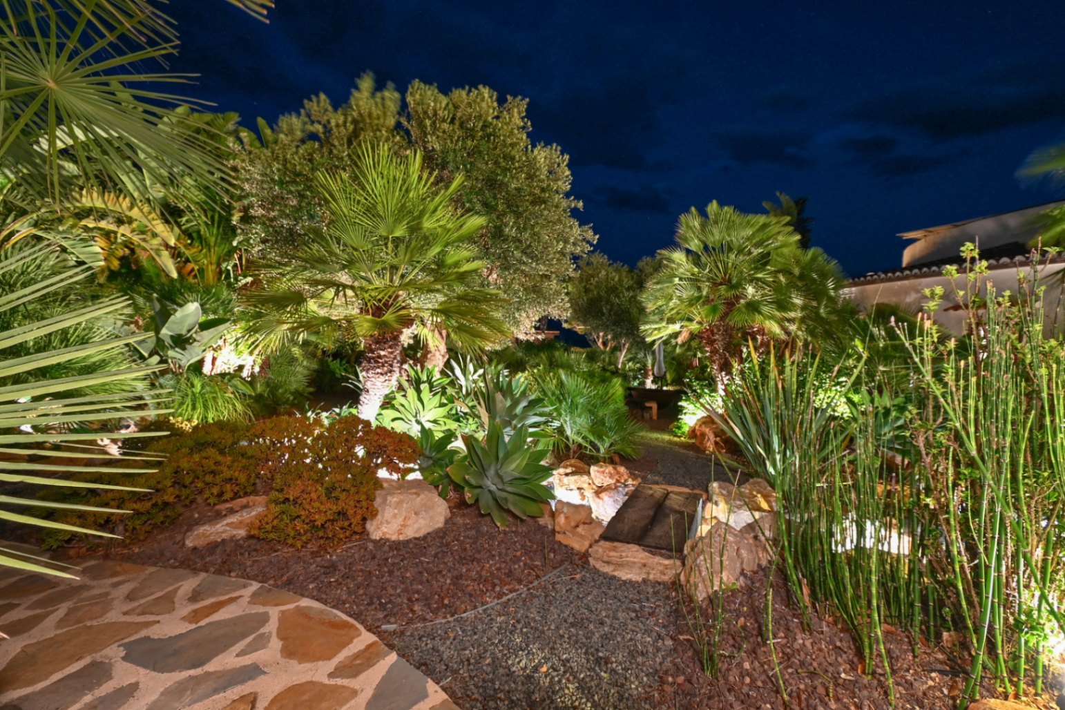 Illuminated Mediterranean garden at night featuring palm trees, agaves, and a gravel path with a small wooden bridge among decorative stones.