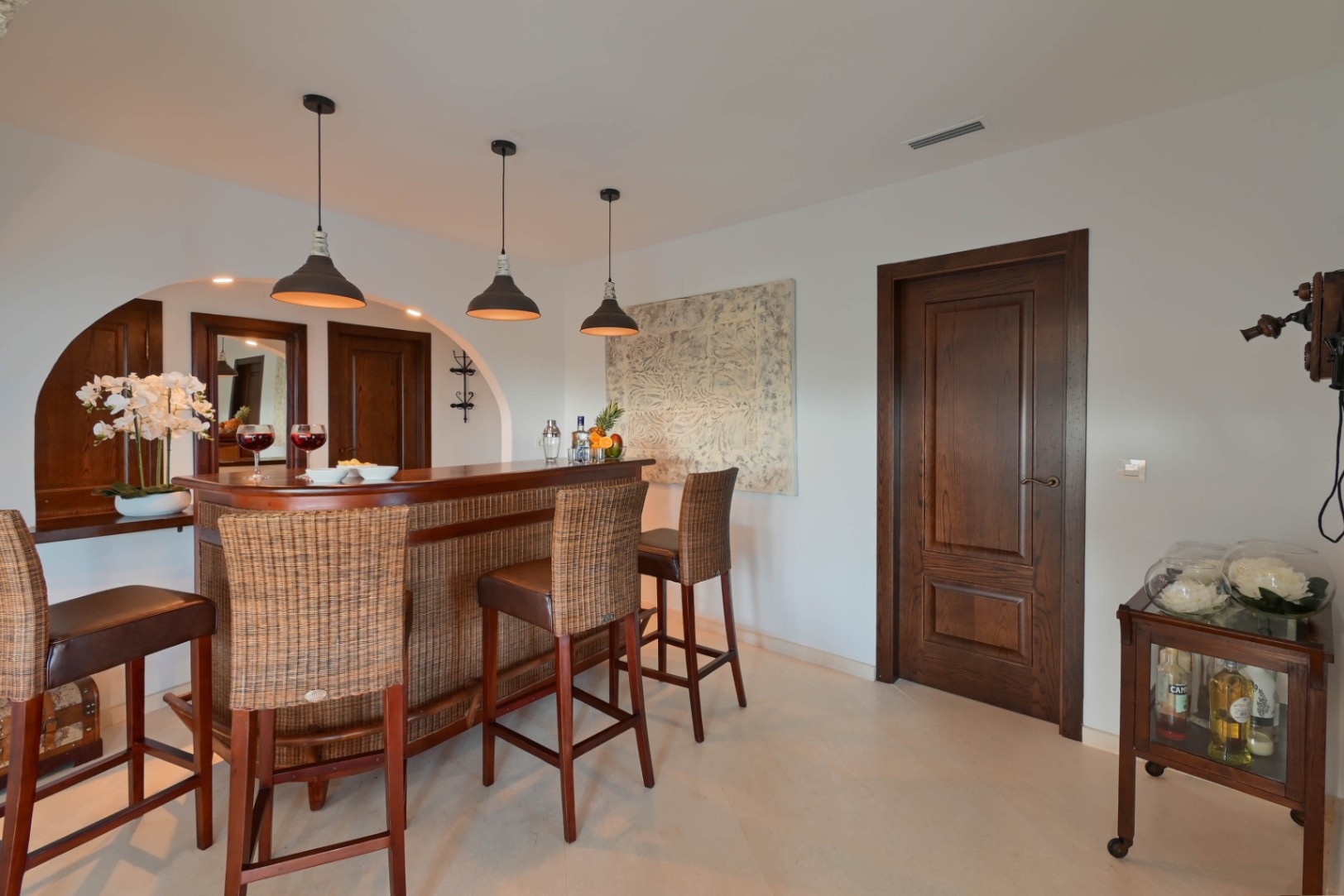 Indoor bar area featuring a curved wooden counter with wicker accents, high-backed stools, and industrial pendant lighting against dark wood doors.