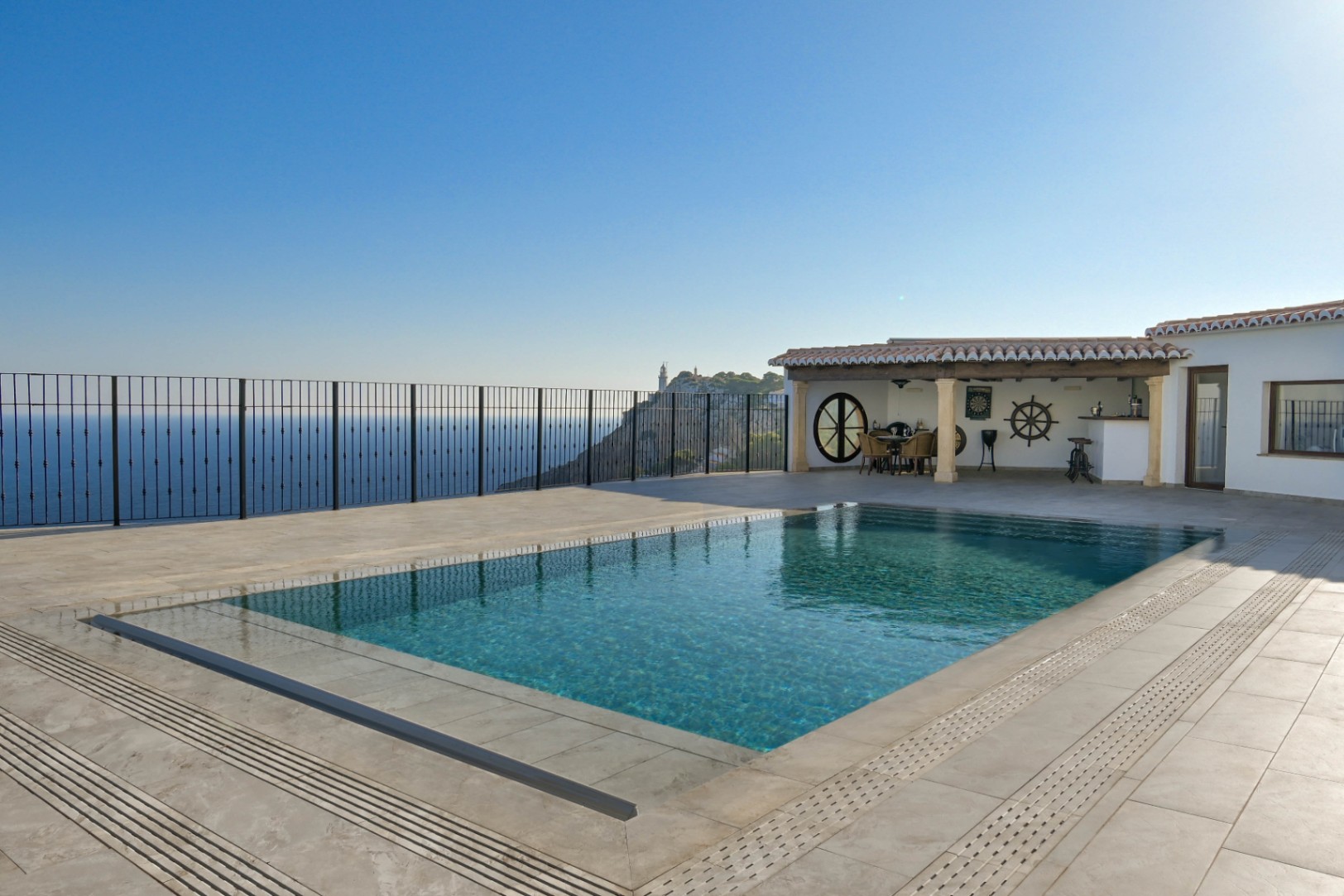 Infinity pool on a stone-tiled terrace with panoramic sea and lighthouse views. Features a covered porch with nautical accents and an outdoor dining area.