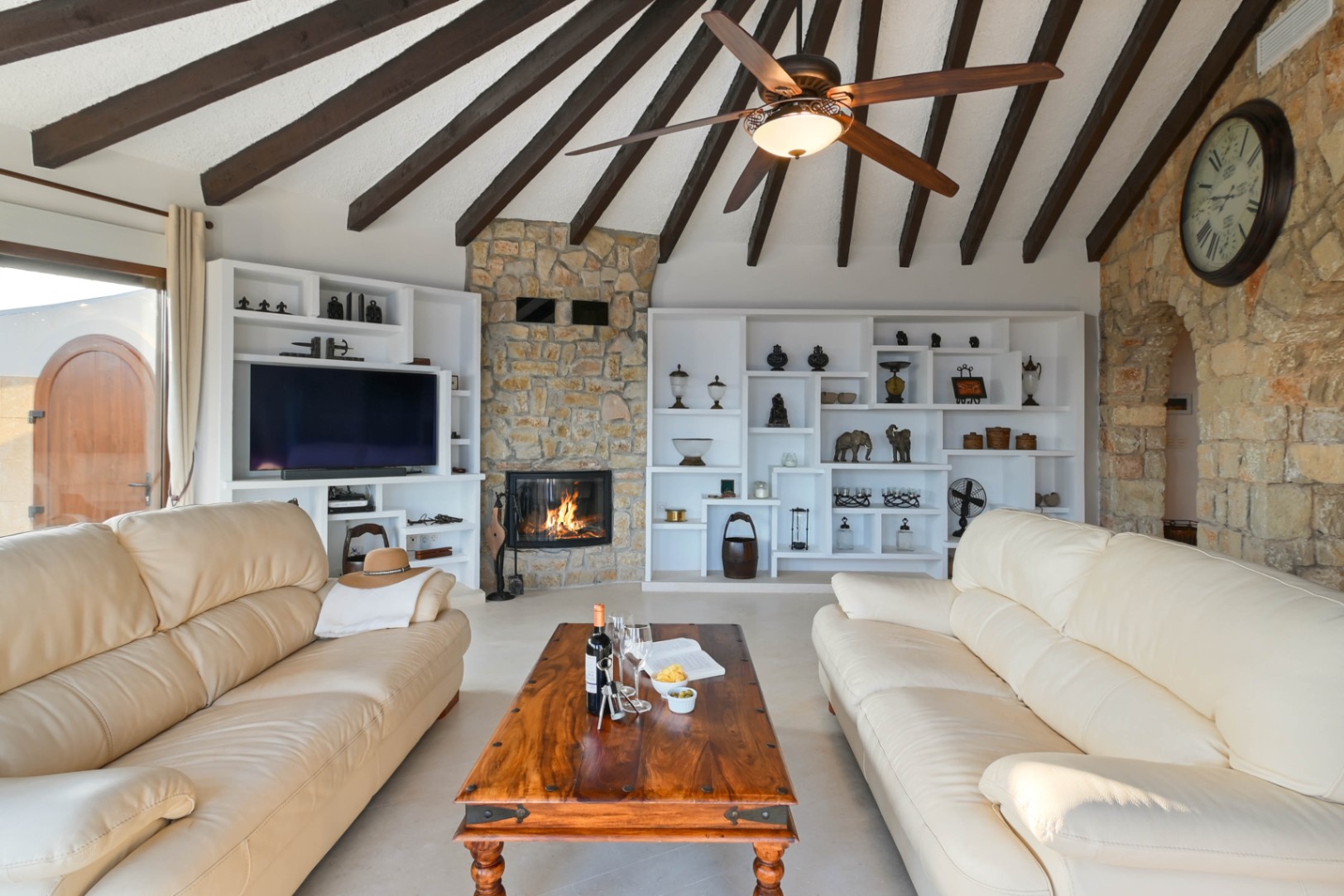 Living room featuring a vaulted ceiling with dark wood beams, a stone fireplace, and custom white shelving. Includes cream leather sofas and a rustic wooden coffee table.