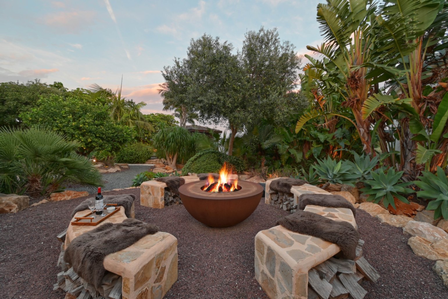 Outdoor circular fire pit area featuring a Corten steel bowl, stone-clad benches with integrated wood storage, and lush tropical landscaping at dusk.