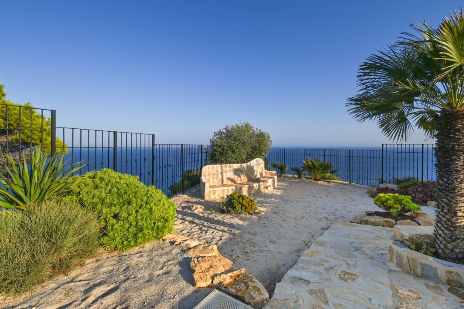 Outdoor viewpoint featuring a built-in stone bench, black metal fencing, and panoramic sea views framed by Mediterranean vegetation and palm trees.
