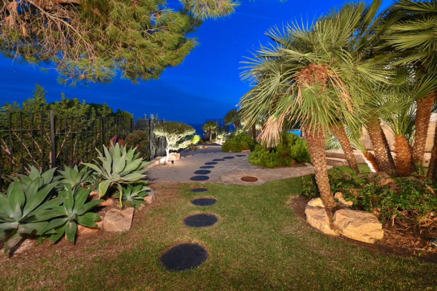 Path of circular stone steps through a Mediterranean garden with palm trees, agaves, and integrated night lighting under a twilight sky.