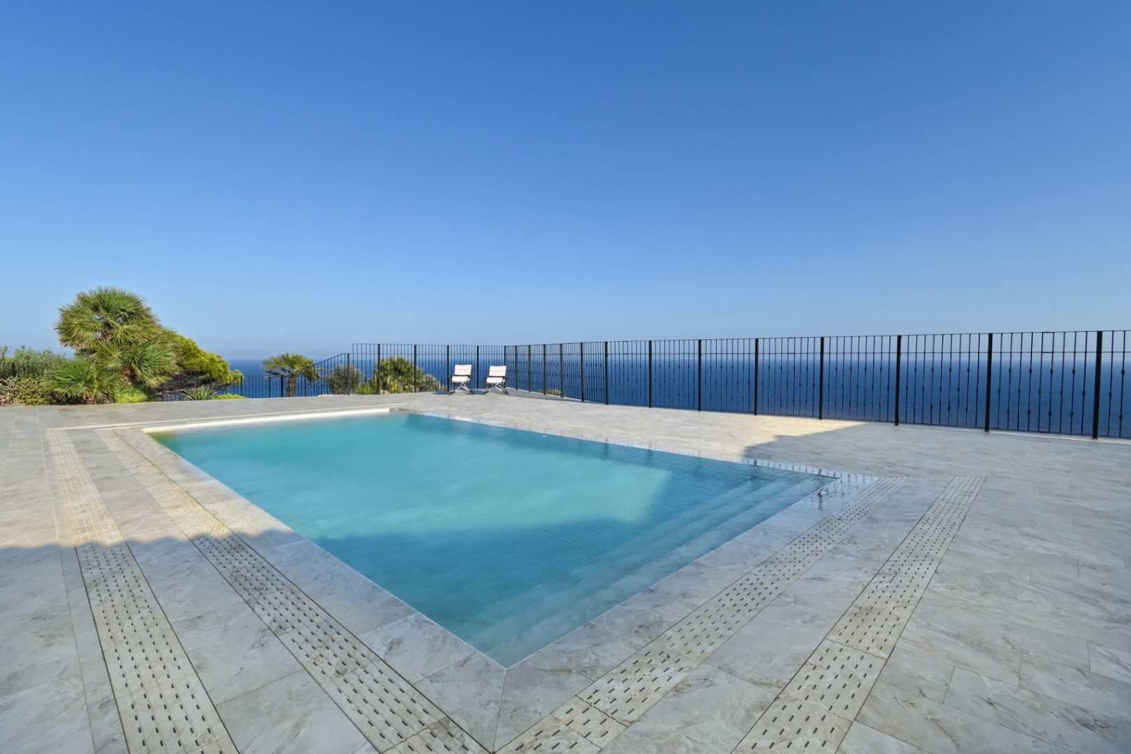 Rectangular swimming pool with integrated steps, set on a light stone terrace with black metal railings and panoramic sea views.