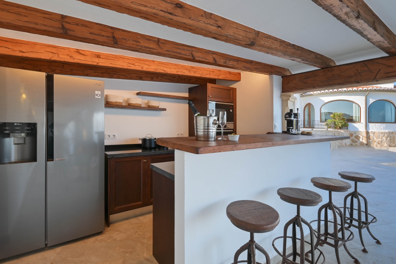 Rustic-modern kitchen bar featuring exposed wooden beams, an LG stainless steel refrigerator, and dark wood cabinetry. A white breakfast bar with industrial stools overlooks the patio.