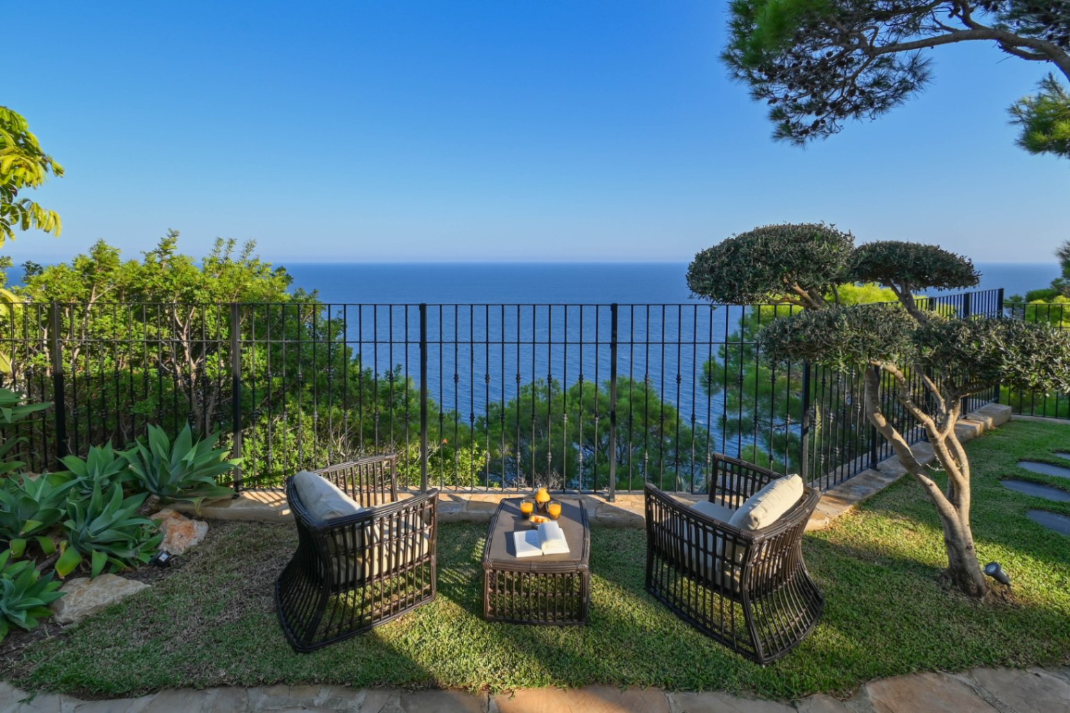 Seating area with dark wicker armchairs and a matching table on a grass lawn, offering panoramic sea views behind a wrought iron railing.