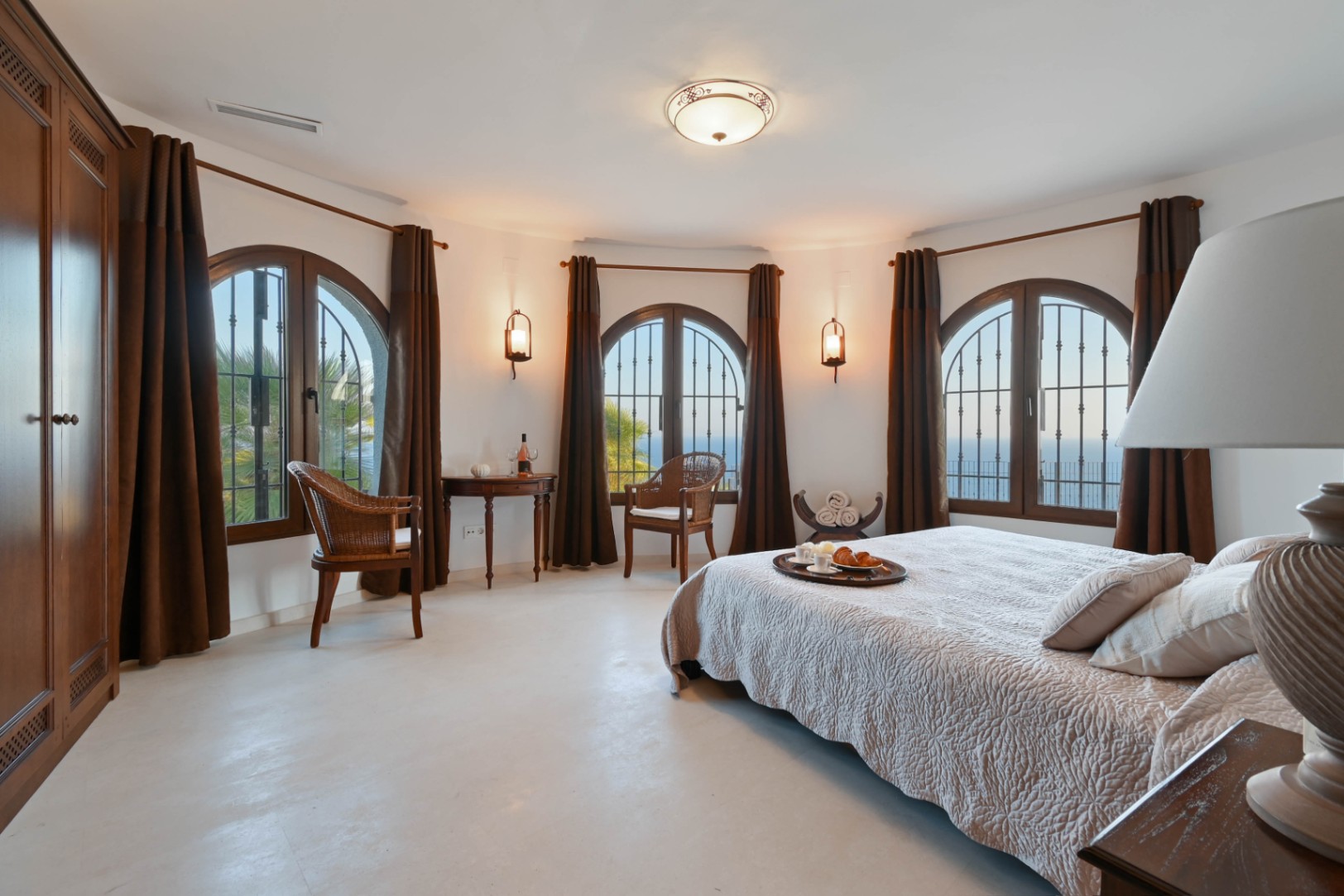 Spacious bedroom featuring a curved wall with three arched windows, dark wood accents, wicker seating, and panoramic sea views.