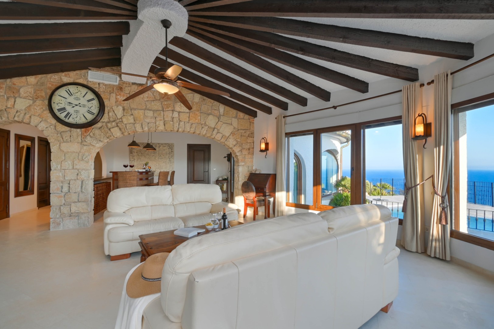 Spacious living area featuring a vaulted ceiling with dark wood radial beams, a natural stone archway, and panoramic sea views through large windows.
