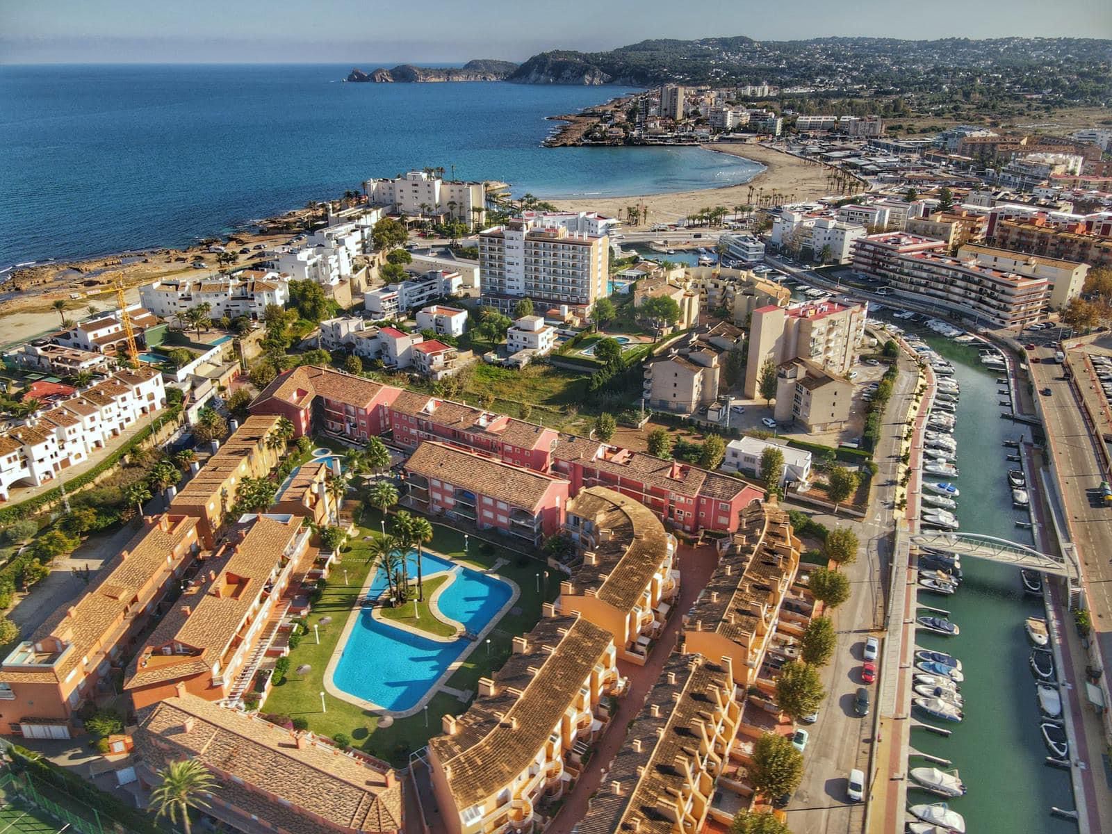 Aerial view of a coastal residential complex featuring a curved swimming pool, terracotta roofs, and proximity to a boat canal and sandy beach.