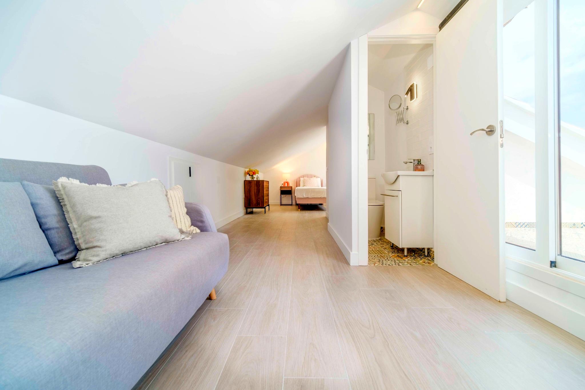 Bright attic suite featuring sloped white ceilings, light wood-effect flooring, a lounge area, and an en-suite bathroom with patterned tiling.