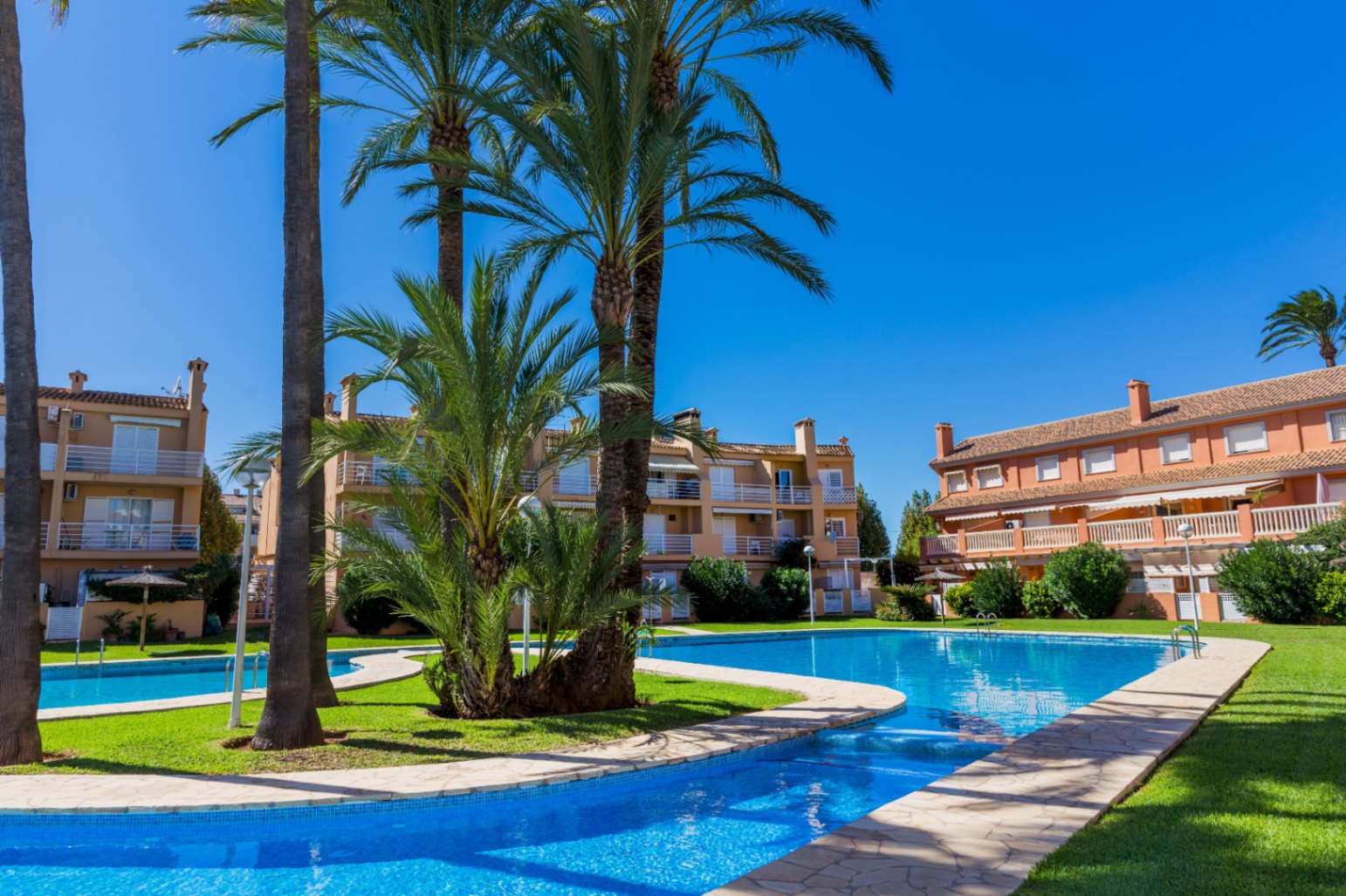 Large communal swimming pool with a connecting channel, surrounded by manicured lawns and palm trees, set against terracotta-colored residential buildings.