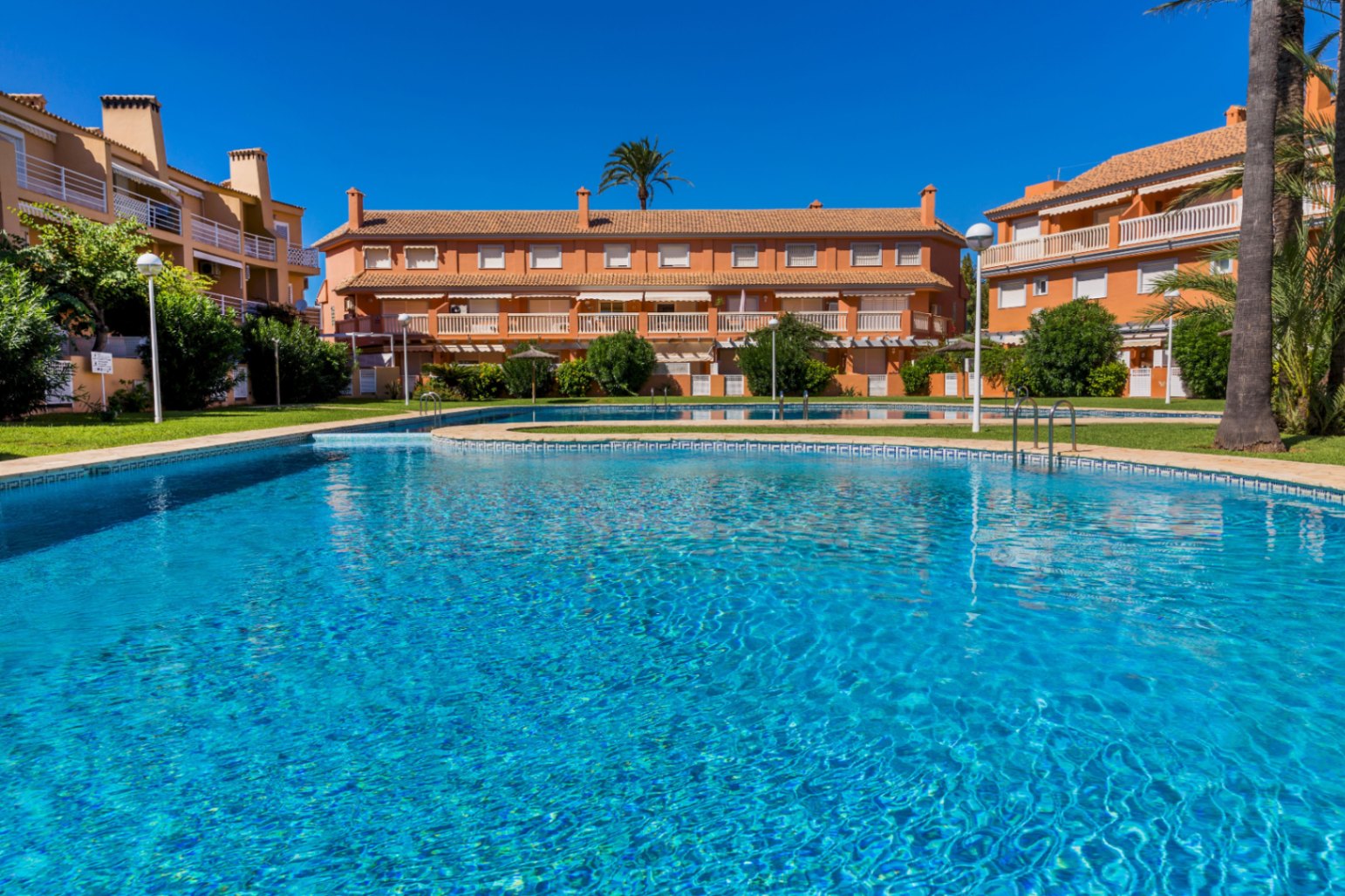 Large communal swimming pool with blue mosaic tiling, surrounded by landscaped gardens and terracotta-colored residential buildings under a clear sky.