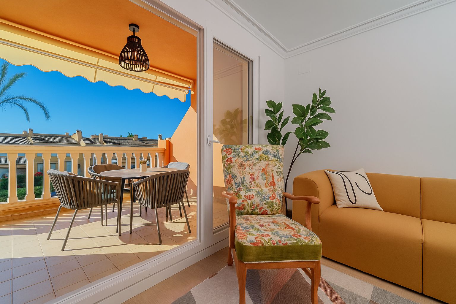 Living area with a mustard sofa and floral armchair opening onto a private terrace with an outdoor dining set, yellow awning, and clear sky views.
