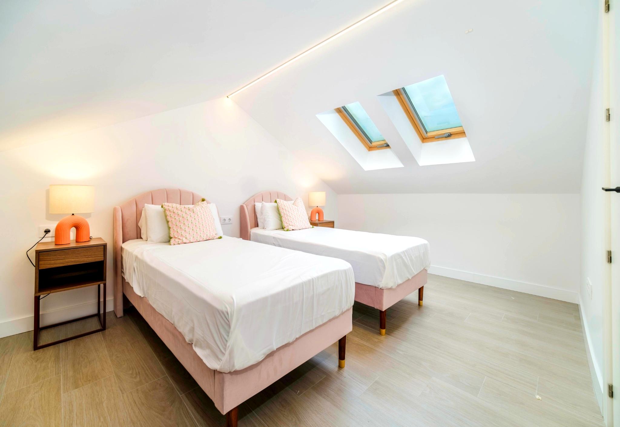 Modern attic bedroom featuring twin beds with pink velvet headboards, dual skylights, and integrated LED ceiling lighting over light wood flooring.