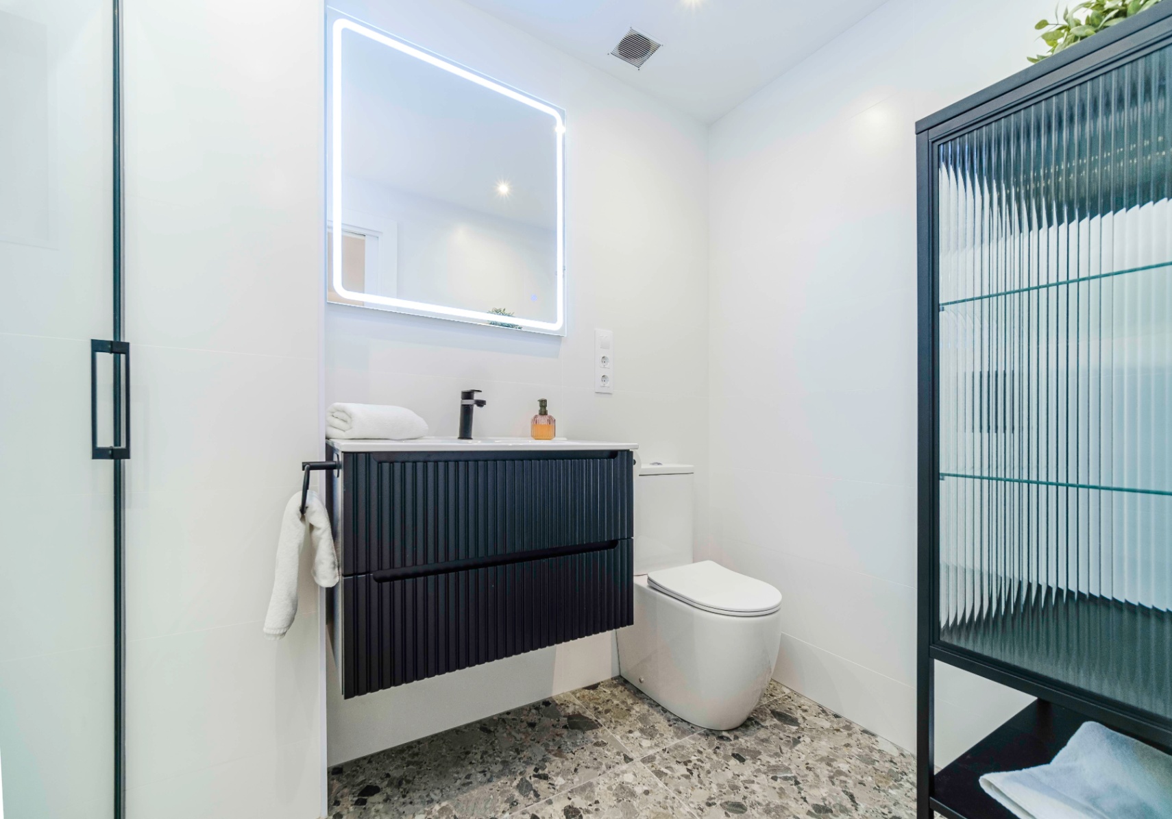 Modern bathroom featuring a black fluted floating vanity, matte black fixtures, and a backlit LED mirror against grey terrazzo flooring.