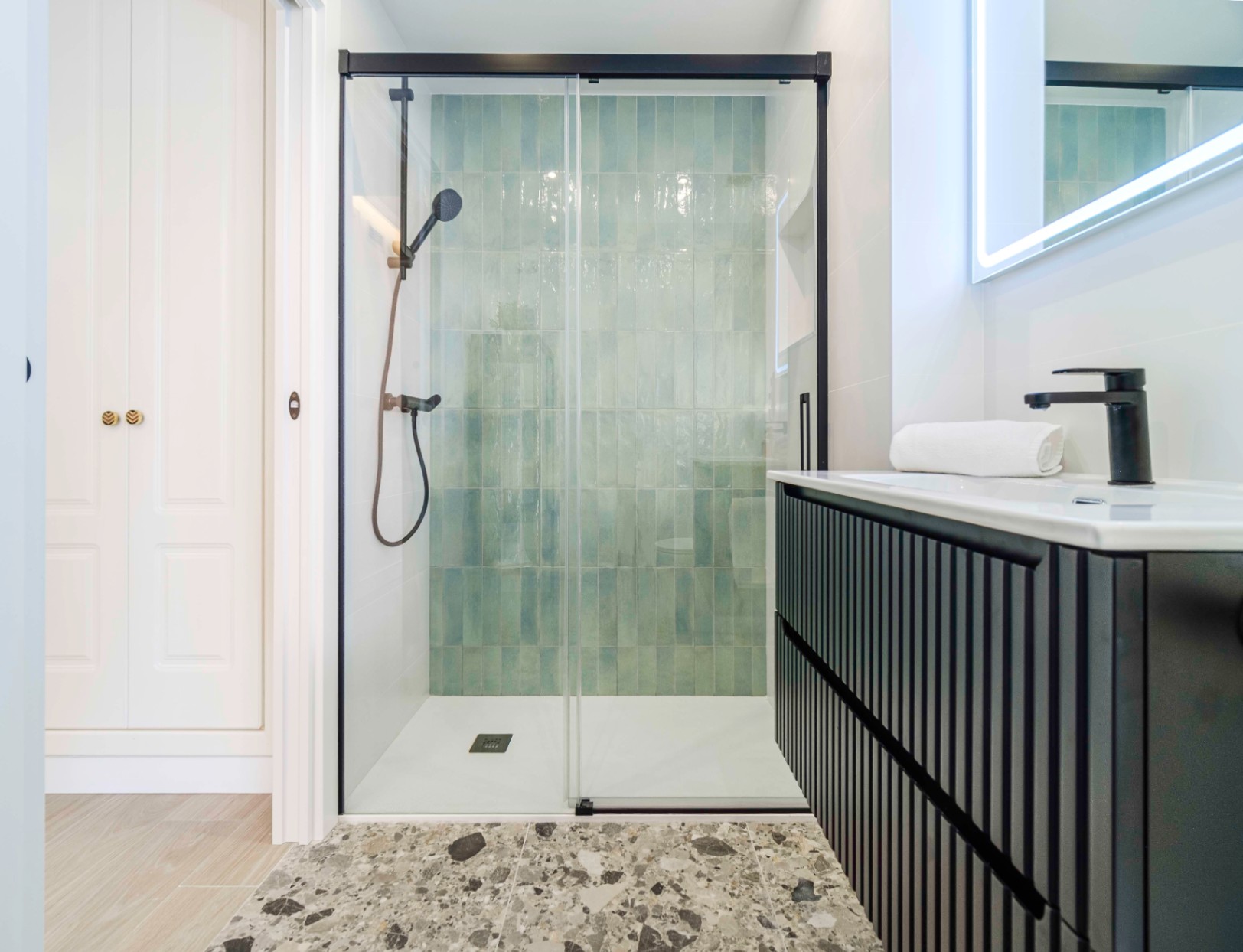 Modern bathroom featuring a walk-in shower with turquoise vertical tiles, black fixtures, and a slatted black vanity with a backlit mirror.