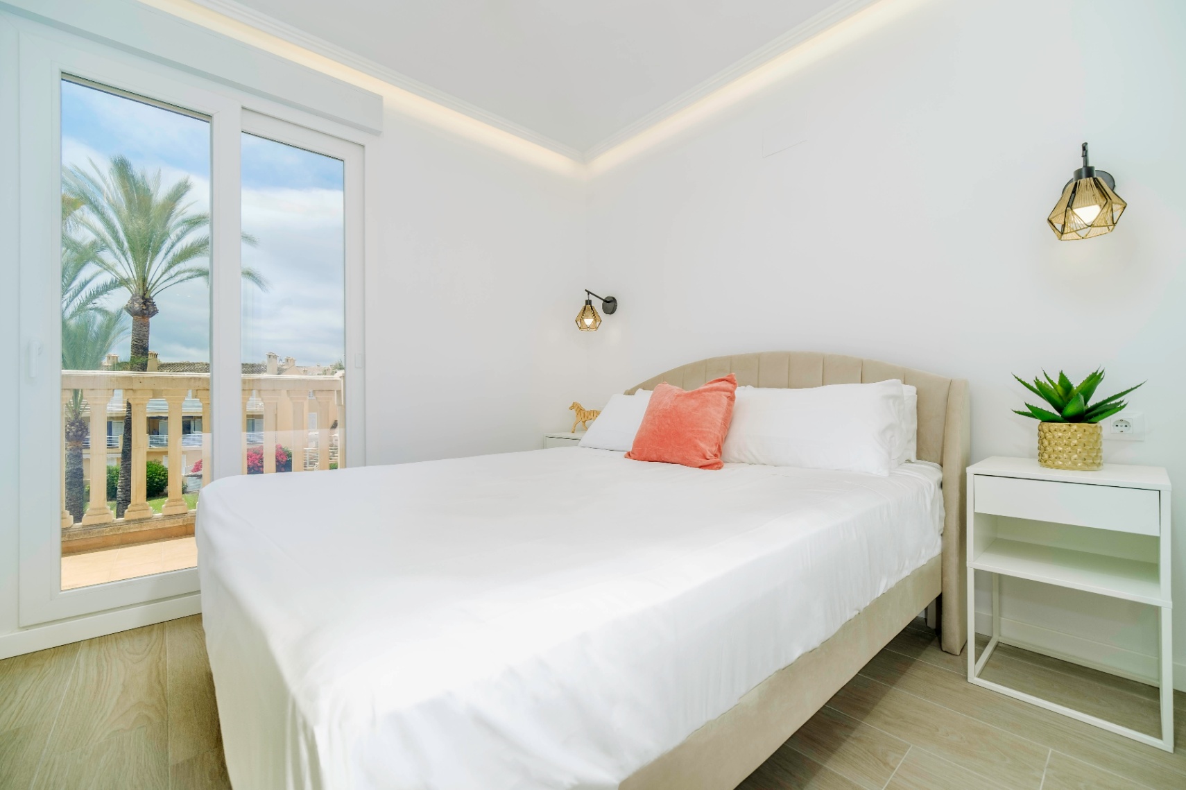 Modern bedroom featuring a beige upholstered bed, geometric wall lamps, and a sliding glass door opening to a terrace with palm tree views.