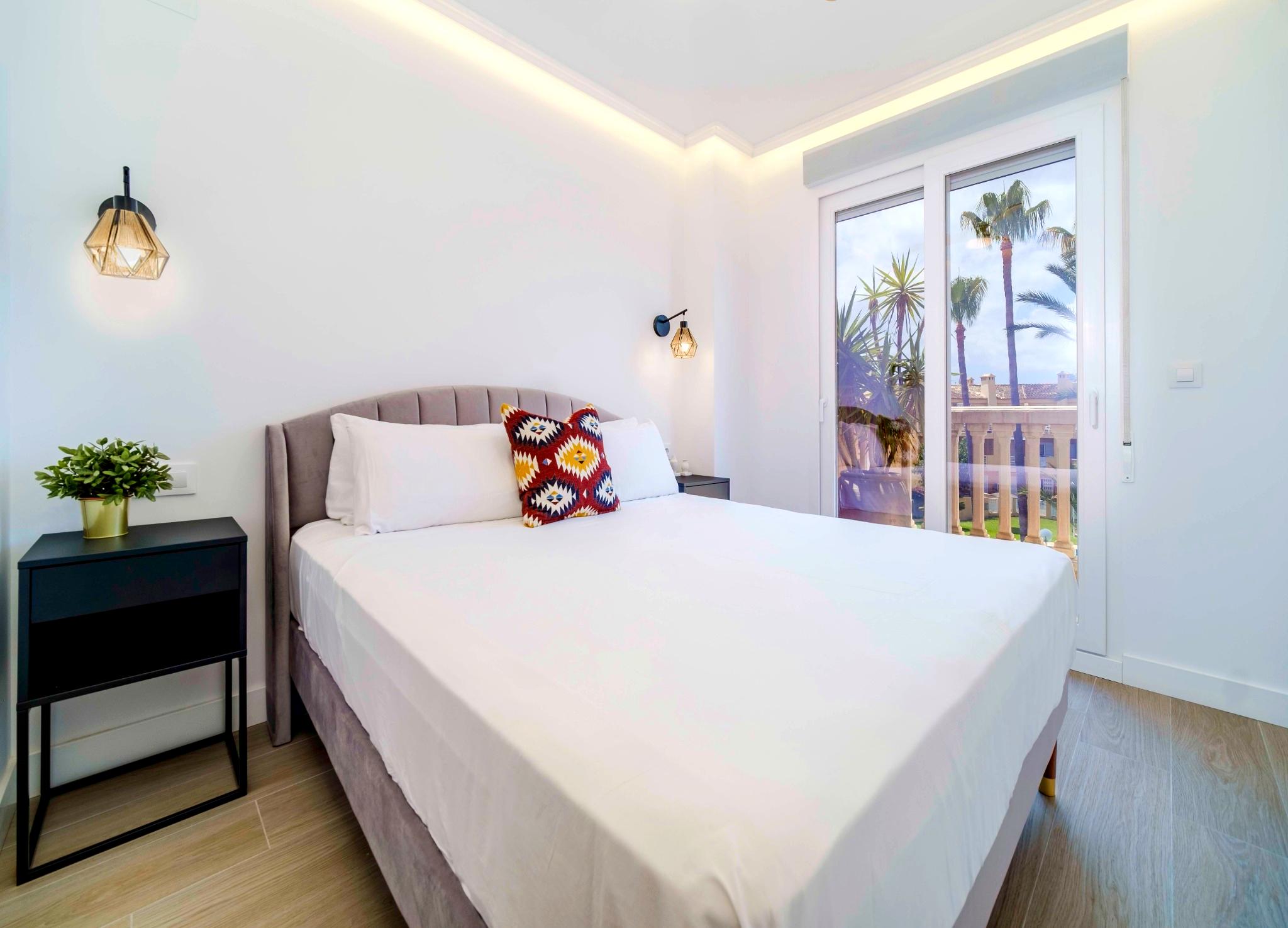 Modern bedroom featuring a velvet headboard, geometric wall sconces, and recessed ceiling lighting. Large sliding doors open to a balcony with views of palm trees.