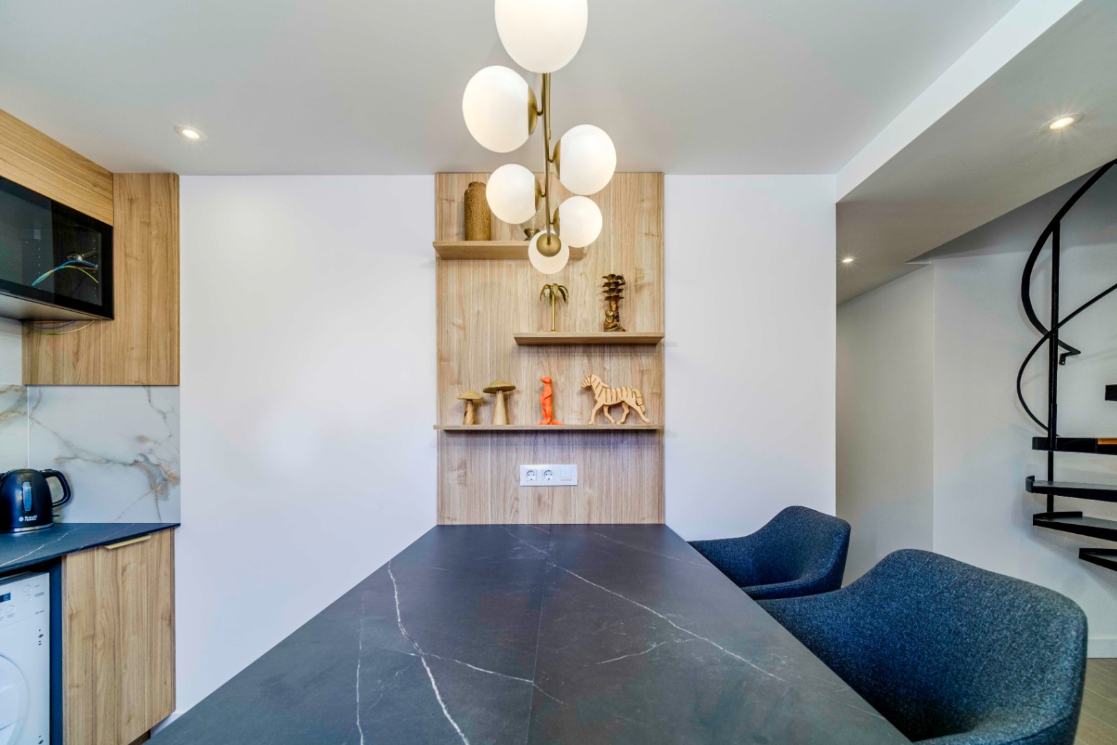 Modern dining area featuring a dark marble-effect table, blue upholstered chairs, and custom wooden shelving illuminated by a brass globe chandelier.