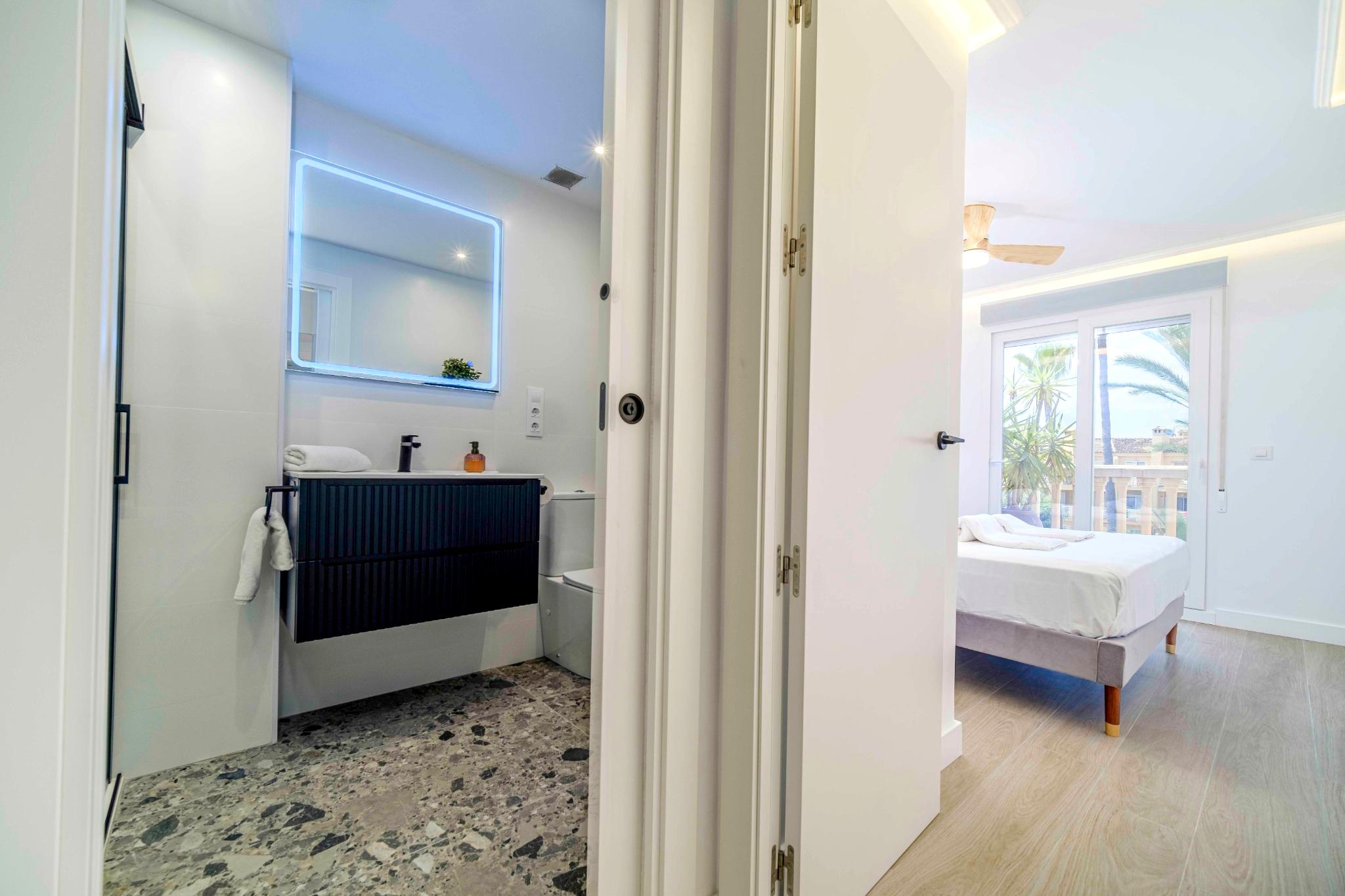 Modern ensuite bathroom with terrazzo flooring and black vanity, adjacent to a bright bedroom featuring a balcony with palm tree views.