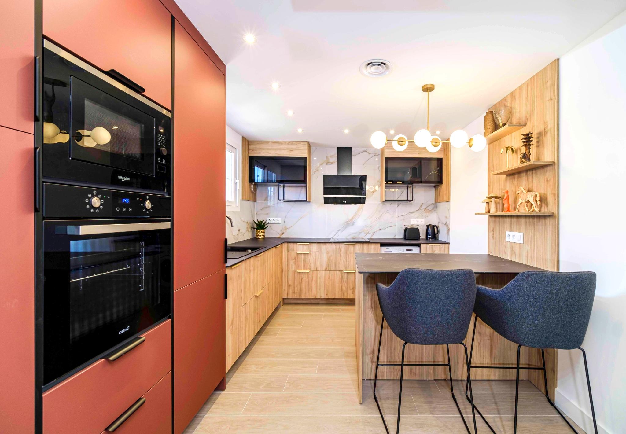 Modern kitchen featuring terracotta tall units, light wood cabinetry, marble-effect backsplash, and a breakfast bar with navy blue stools and a gold globe chandelier.