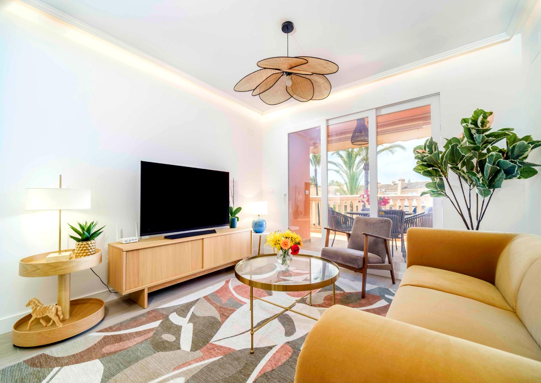 Modern living room featuring a yellow sofa, a petal-shaped wicker pendant light, and large glass doors opening onto a terrace with palm tree views.