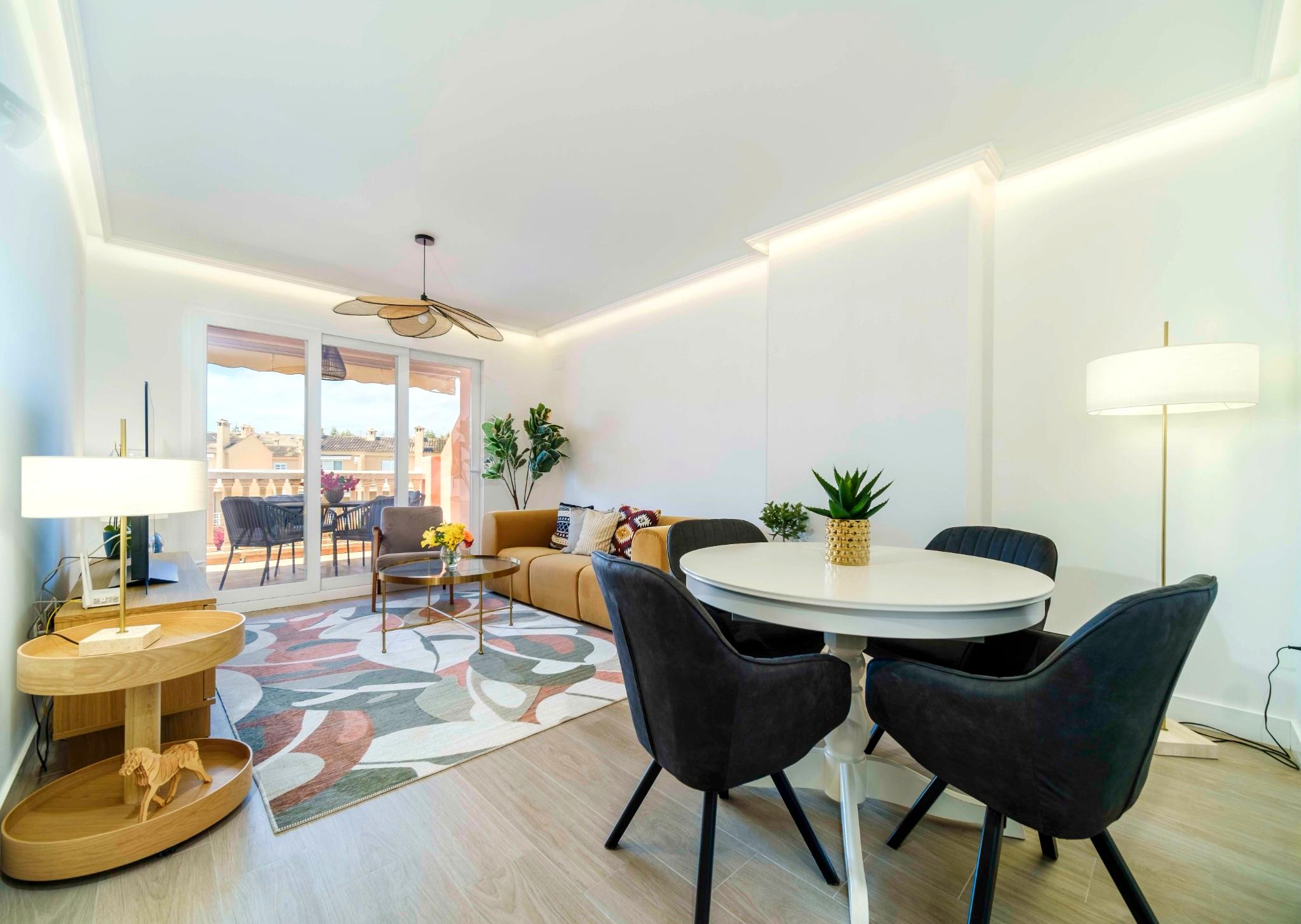 Open-plan living area featuring a round white dining table, mustard sofa, and recessed LED lighting. Sliding glass doors open to a furnished terrace.