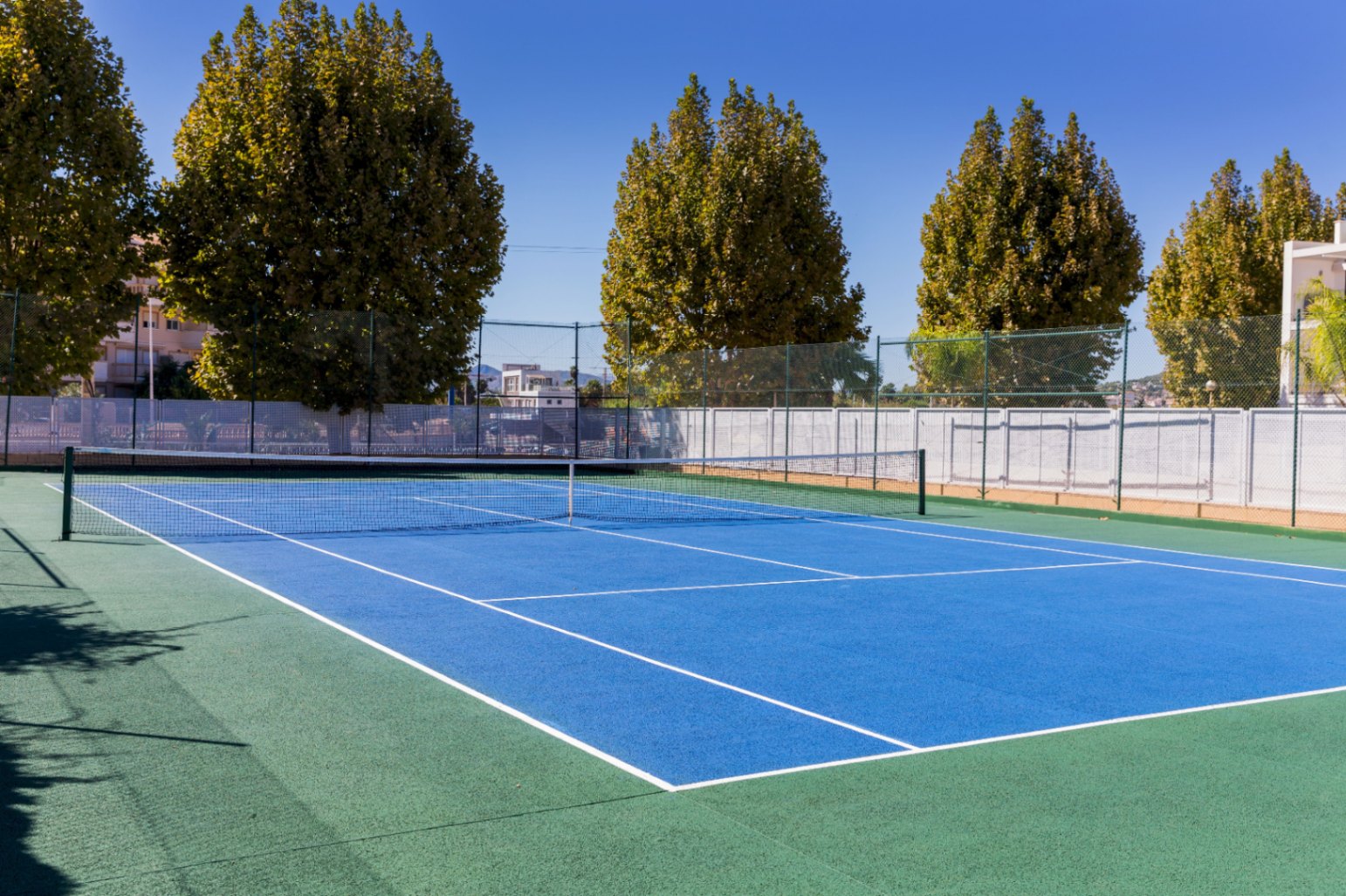 Outdoor hard-surface tennis court with a blue playing area and green perimeter, featuring high fencing and surrounded by mature trees under a clear sky.