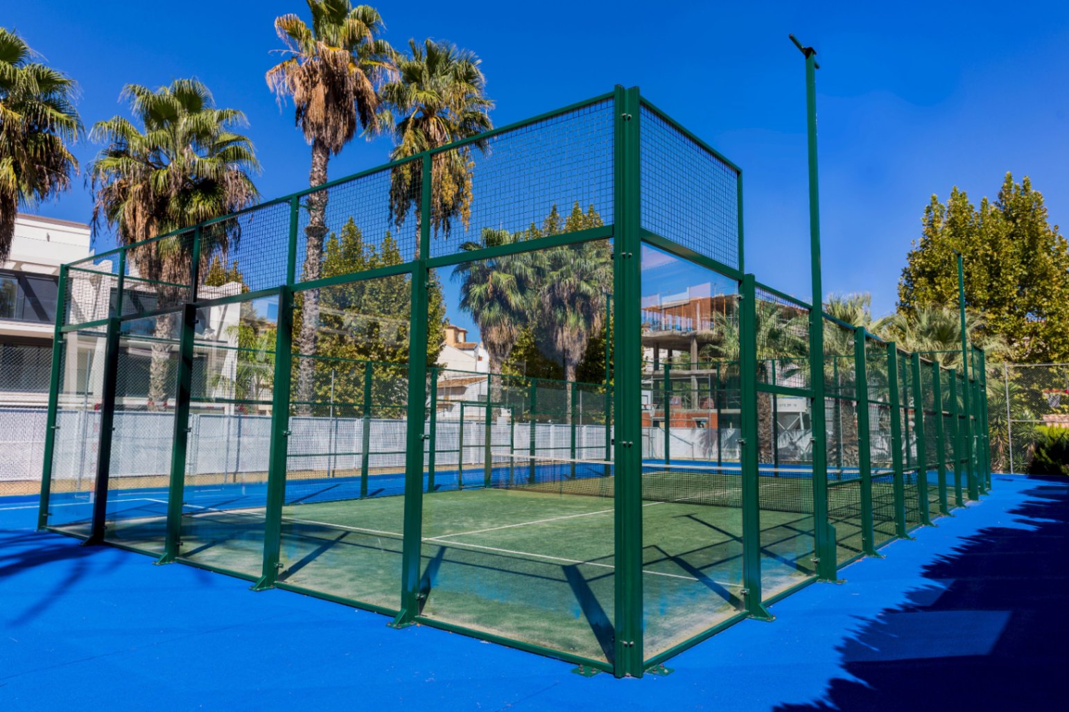 Outdoor paddle tennis court featuring green synthetic turf, tempered glass walls, and a green metal frame, surrounded by palm trees under a clear sky.