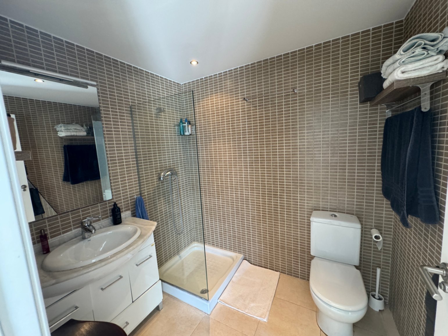 Bathroom with a glass walk-in shower, brown mosaic tile walls, and a white vanity unit featuring a marble countertop and recessed lighting.