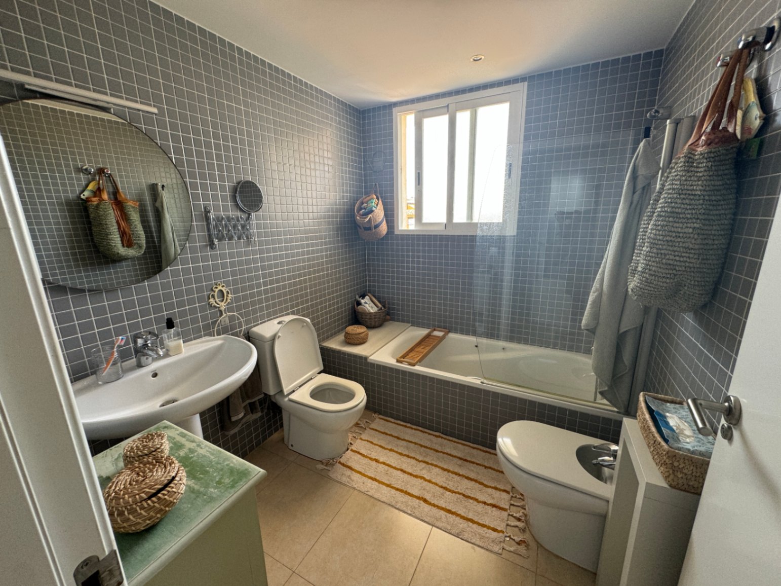 Bathroom with grey mosaic tiling, featuring a white pedestal sink, circular mirror, and a bathtub with a glass screen and natural light from a window.
