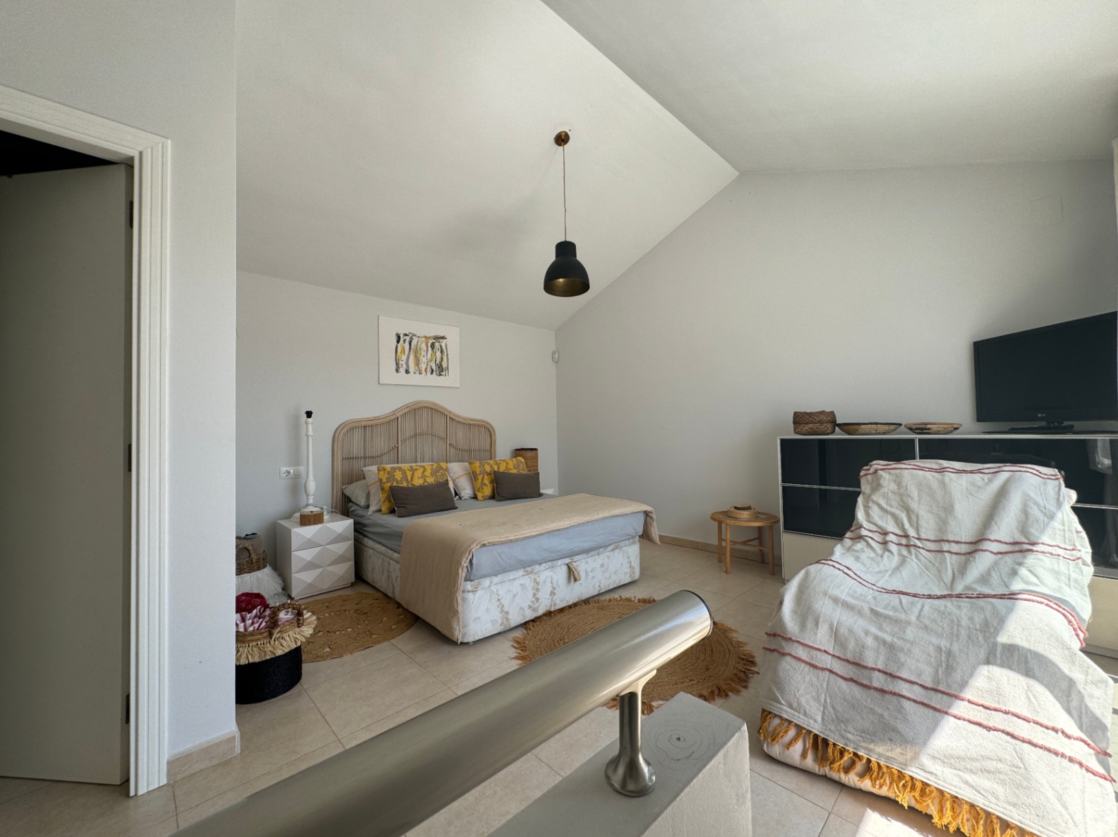 Bedroom with vaulted ceiling, featuring a wicker headboard, black pendant lighting, and tiled flooring. A metal railing overlooks the space from an upper level.