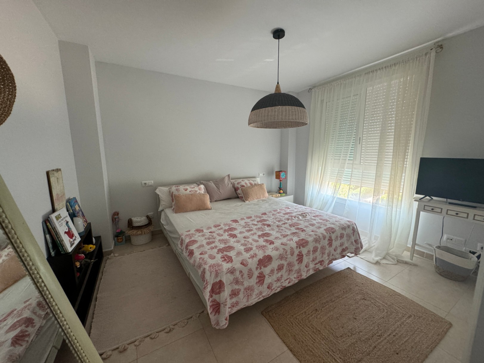 Bright bedroom featuring a double bed with coral-patterned linens, a woven pendant light, and a large window with sheer curtains providing natural light.