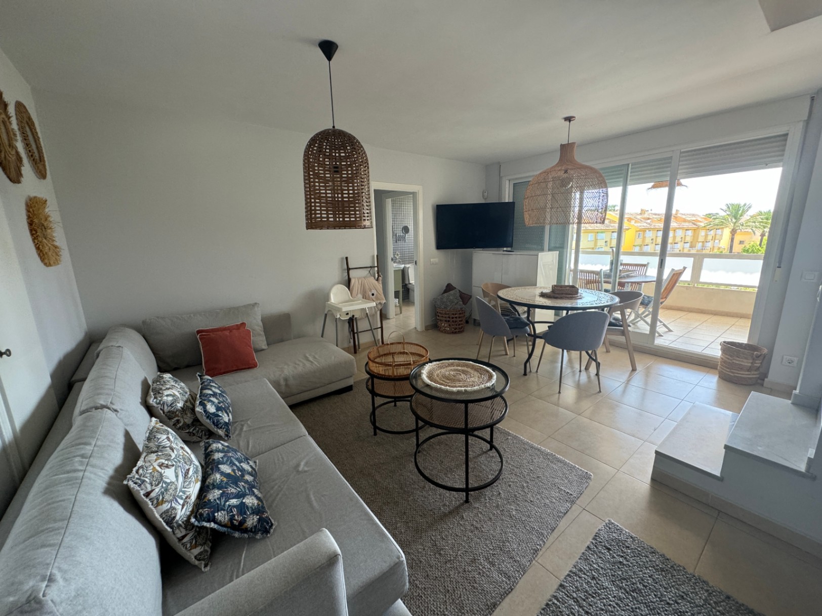 Bright living area featuring a grey sectional sofa, rattan pendant lighting, and a dining set. Sliding doors open to a private terrace with outdoor seating.