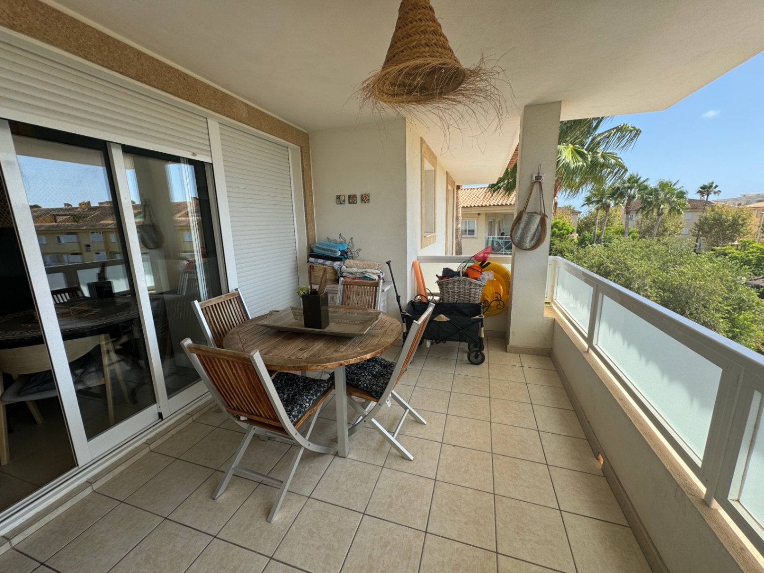 Covered terrace featuring a round wooden dining set, tiled flooring, and a glass railing overlooking palm trees and residential buildings.