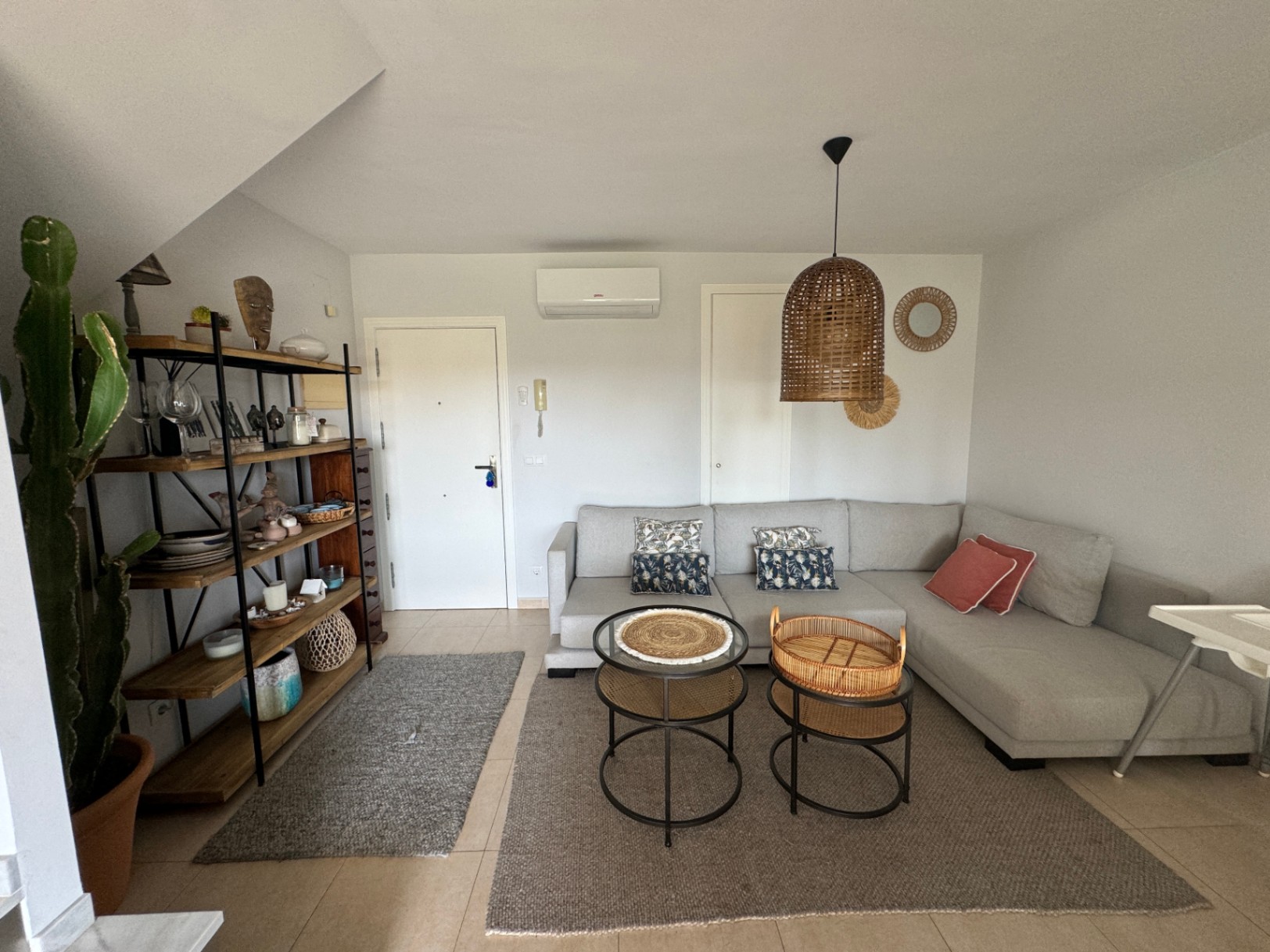 Living area featuring a grey L-shaped sofa, woven rattan pendant lighting, and industrial-style wooden shelving. Tiled floors and neutral tones create a cohesive space.