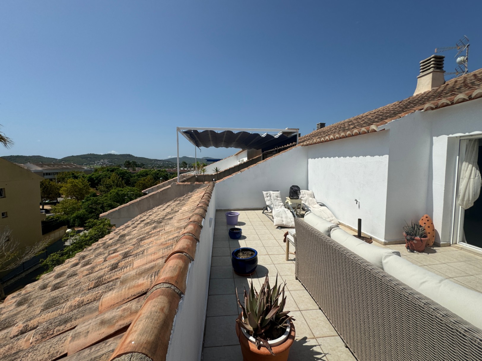 Long rooftop terrace featuring a grey wicker sofa, white sun loungers, and terracotta tiling, offering views of the surrounding green hills and clear sky.