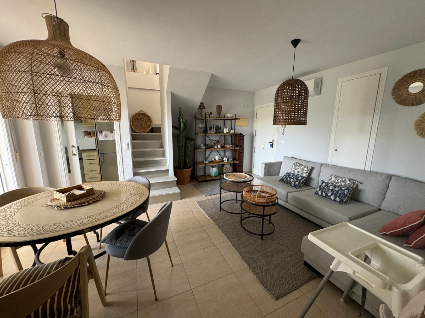 Open-plan living space featuring a grey sectional sofa, rattan pendant lighting, and a dining area with a carved table, all set against neutral tiled flooring.