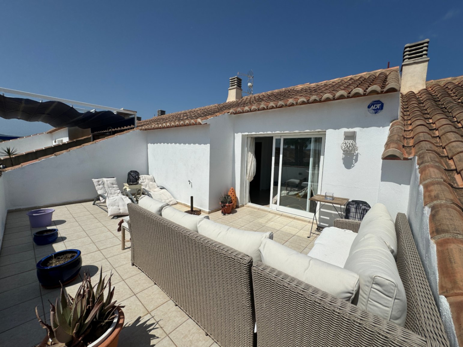 Private rooftop terrace featuring wicker lounge seating, tiled flooring, and sliding glass door access, framed by traditional terracotta roof tiles.