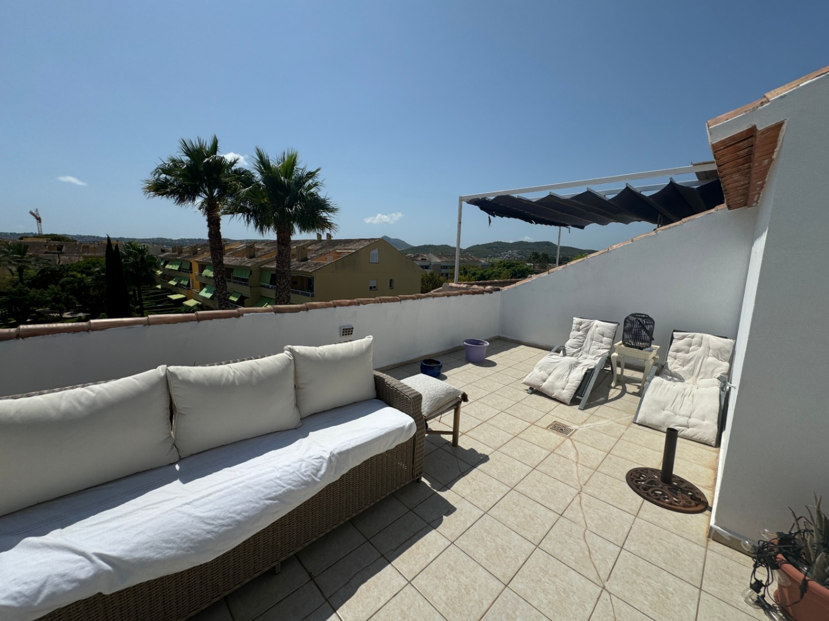 Rooftop terrace with tiled flooring, featuring a wicker sofa, sun loungers, and a retractable awning. Offers views of palm trees and the surrounding neighborhood.