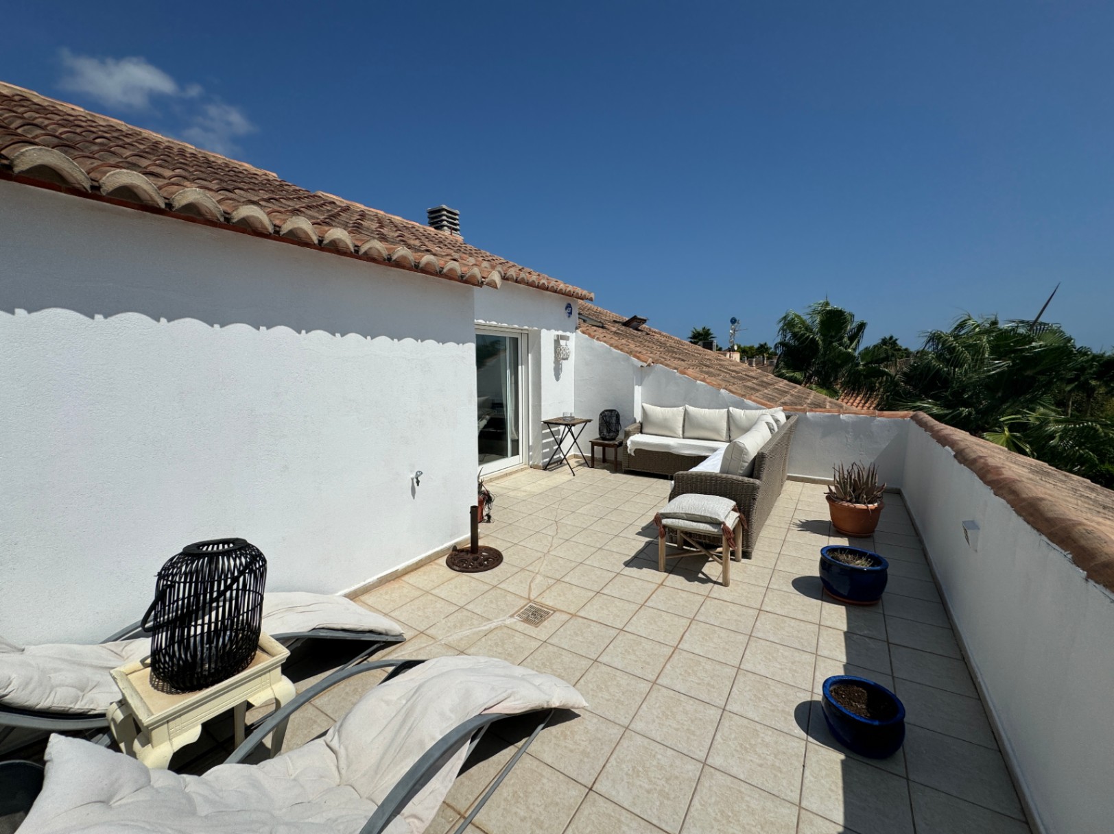 Rooftop terrace with white stucco walls, terracotta tile roofing, a wicker sectional sofa, and tiled flooring overlooking palm trees.