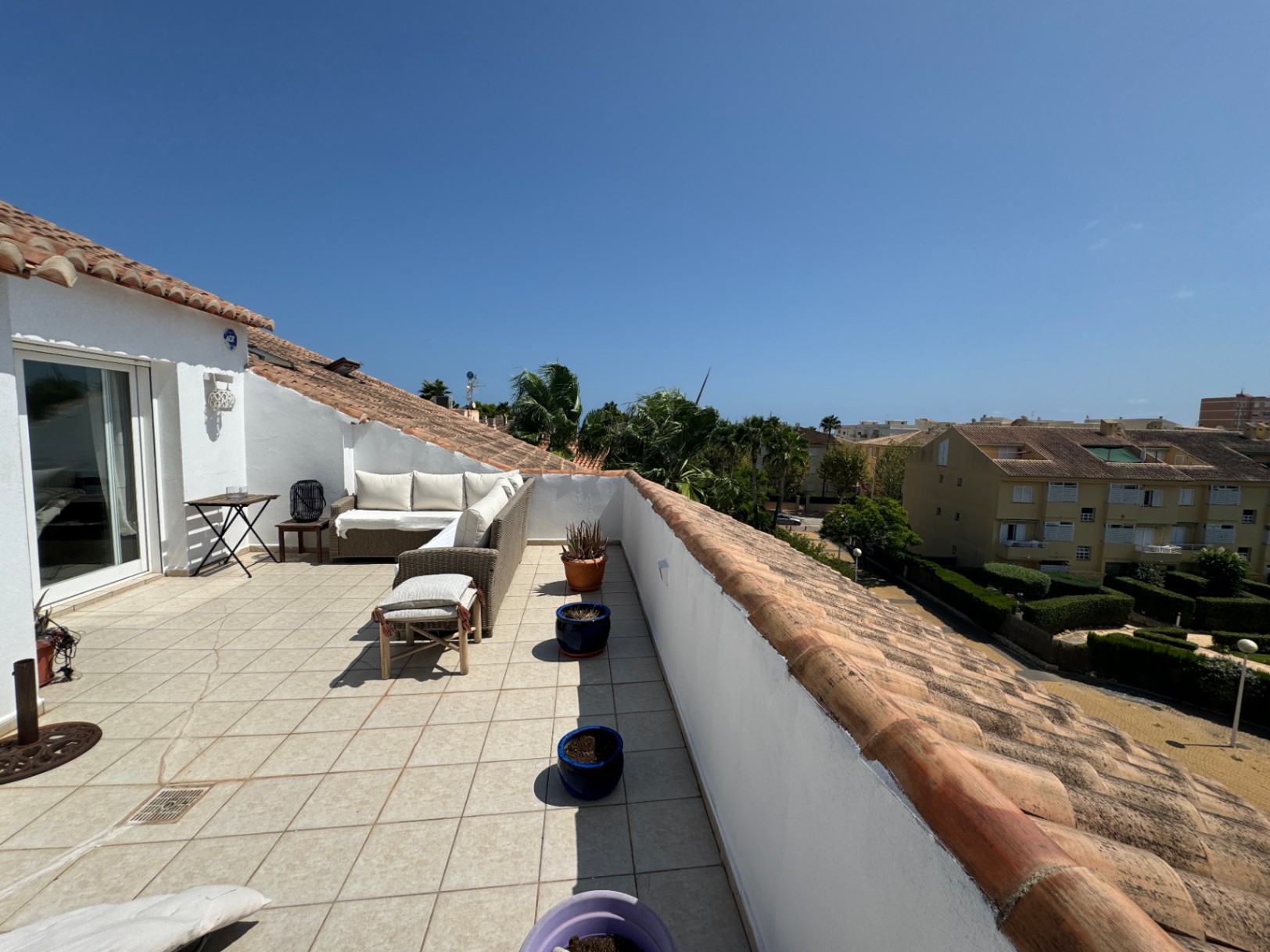 Spacious rooftop terrace featuring light tiled flooring, an L-shaped wicker sofa with white cushions, and views over a residential area with palm trees.