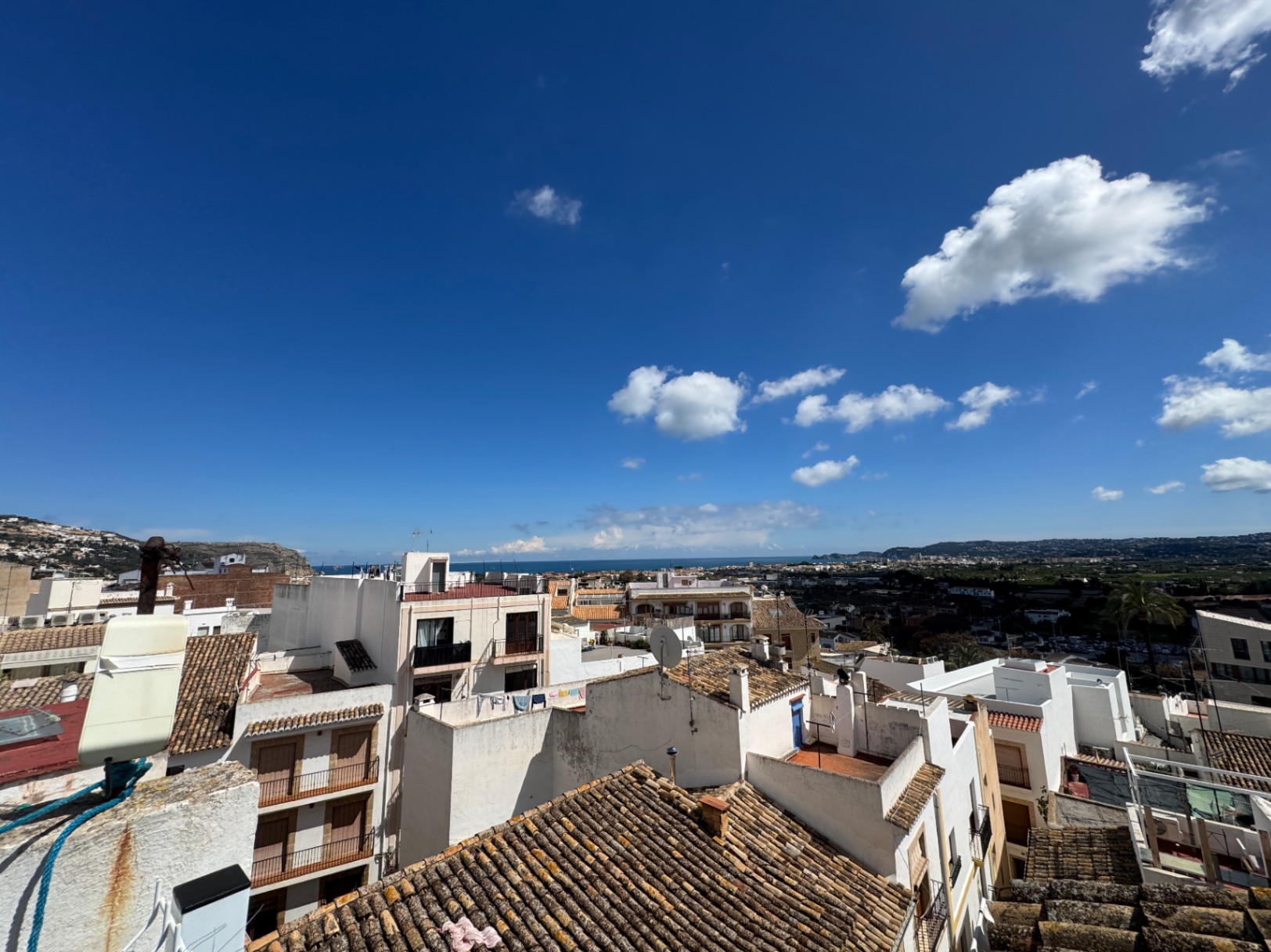 Elevated panoramic view over traditional white-washed buildings and terracotta tiled roofs towards the Mediterranean Sea under a clear blue sky.