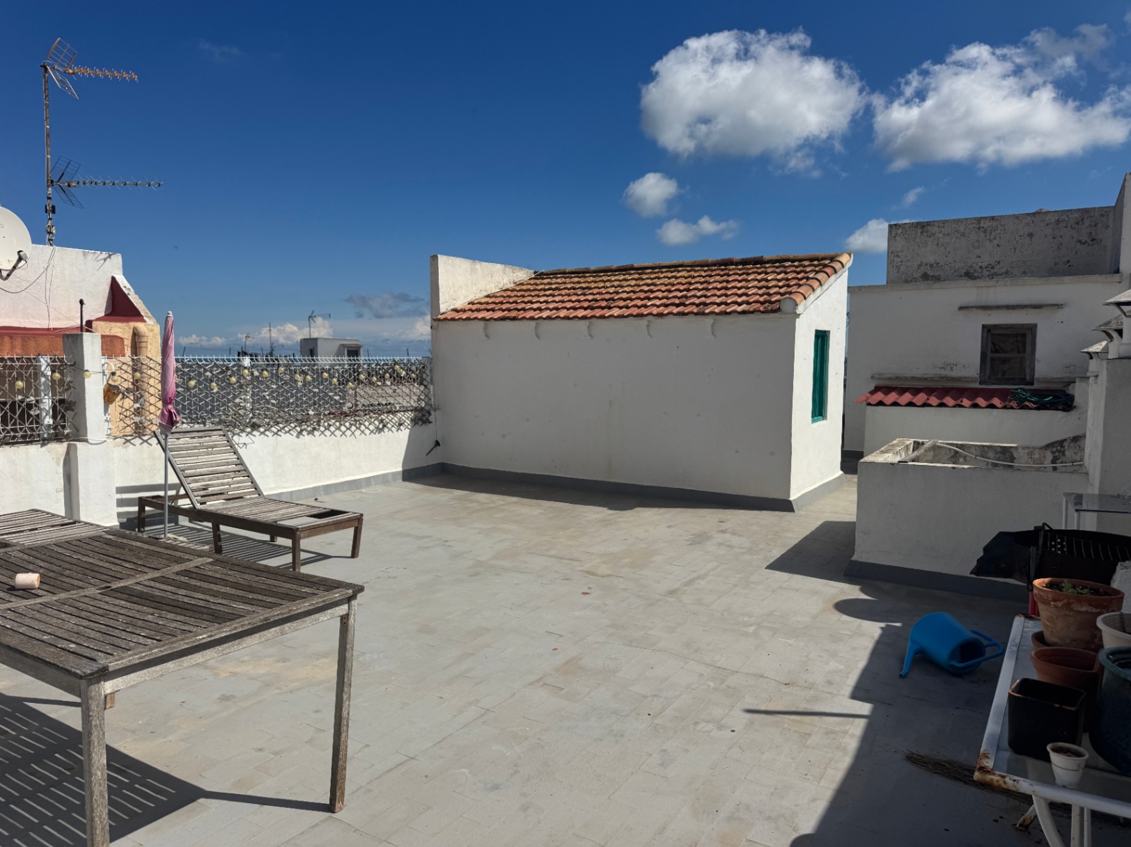 Rooftop terrace featuring tiled flooring, weathered wooden furniture, and a white outbuilding with a terracotta roof under a clear blue sky.