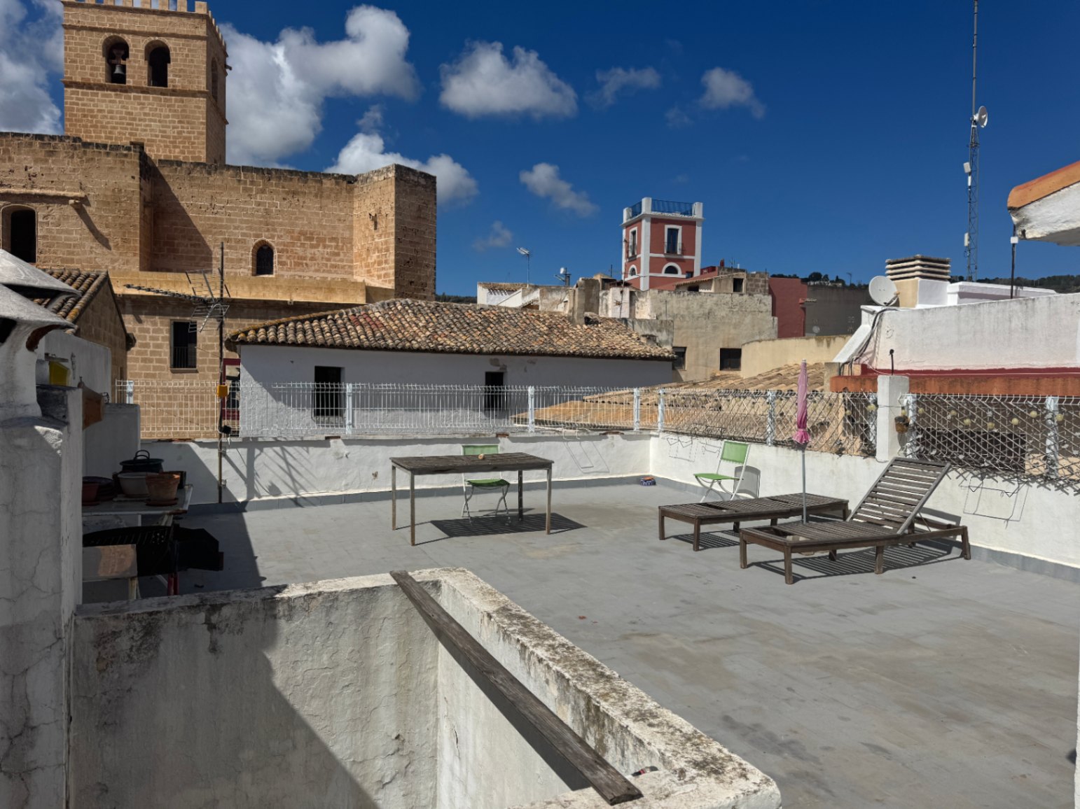 Rooftop terrace featuring wooden sun loungers, a dining table, and views of a historic stone tower and surrounding village rooftops.